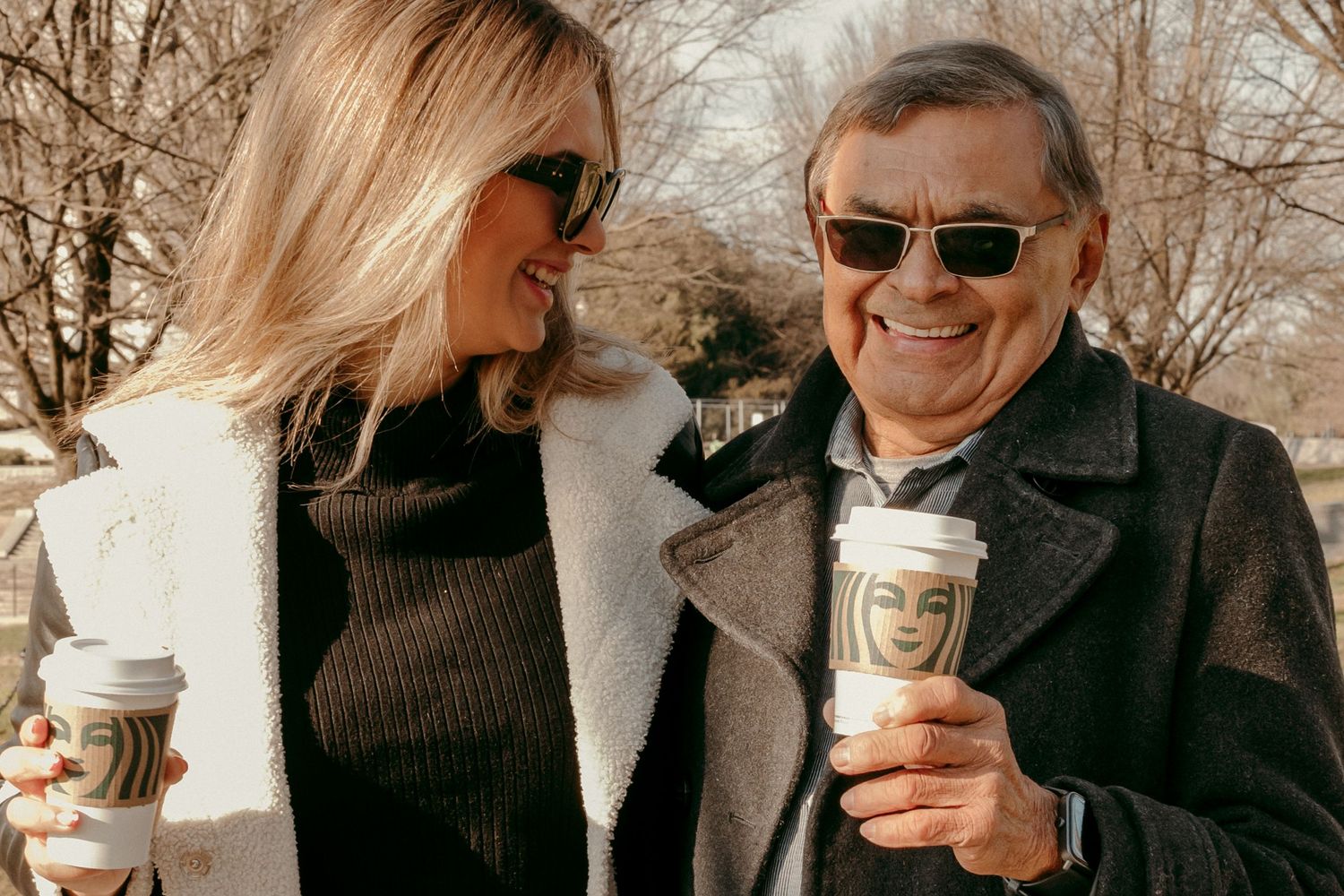 A pair sharing coffee and laughter while sitting outdoors in winter coats.