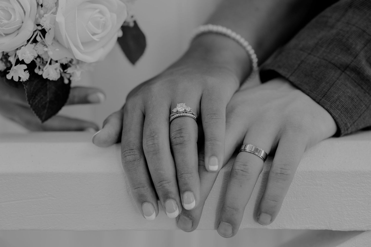 Black and white photo showing two hands wearing wedding rings resting together on a light surface beside roses.