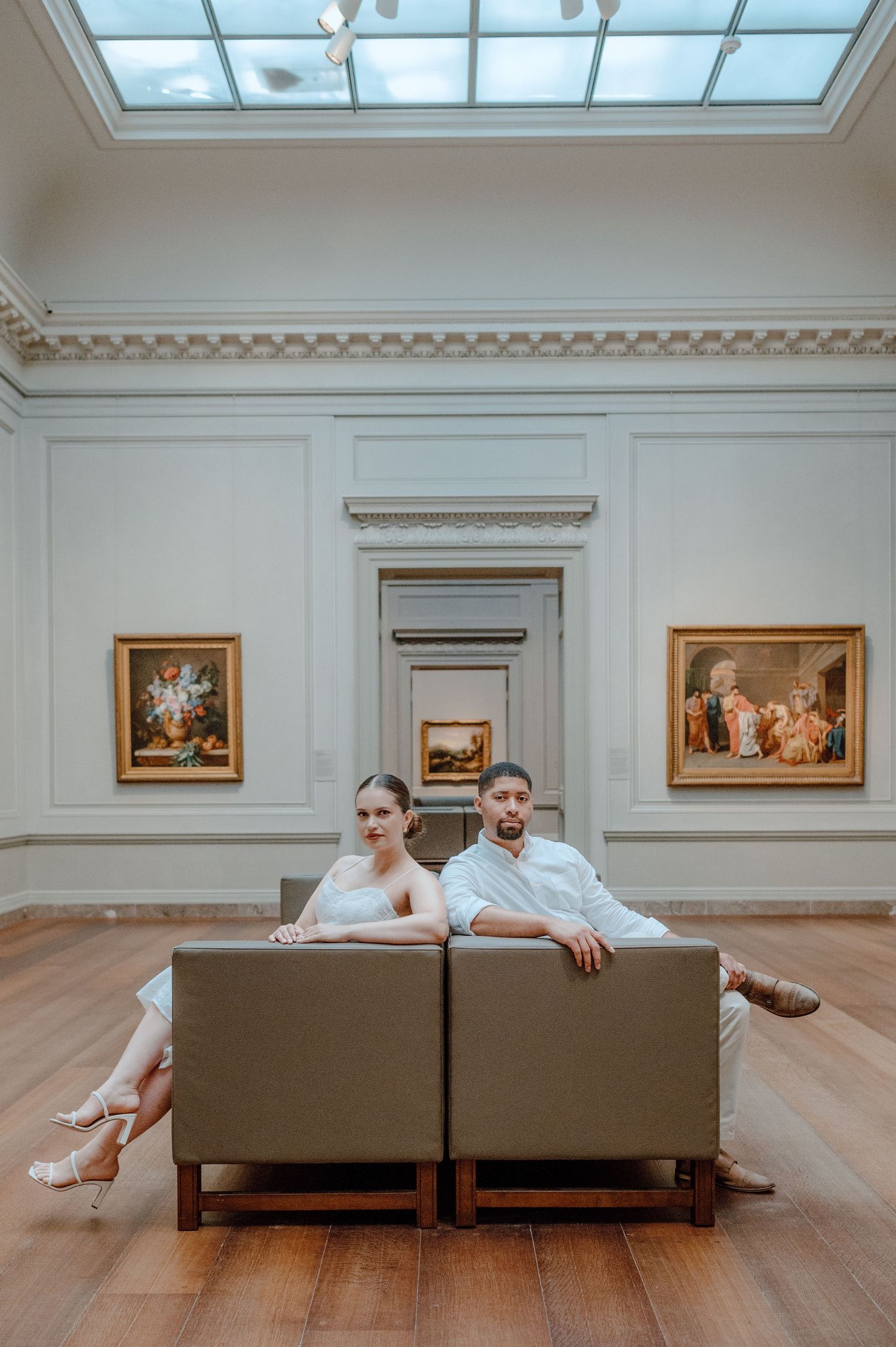A couple relaxes on a brown couch in an elegant museum gallery with artwork and a skylight above.