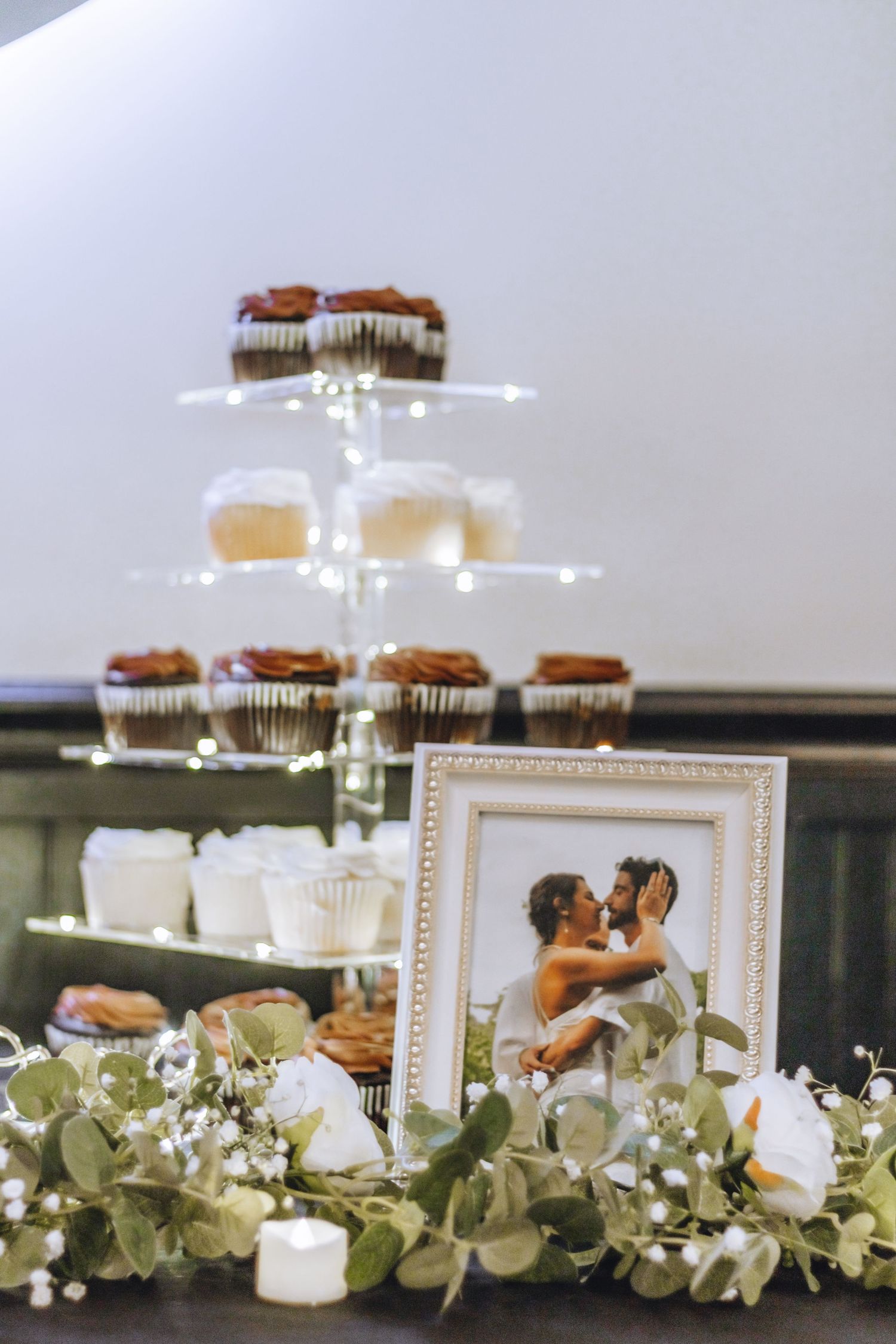 Wedding dessert table display with tiered cupcake stand, floral decorations, and a framed photo on an elegant green surface.