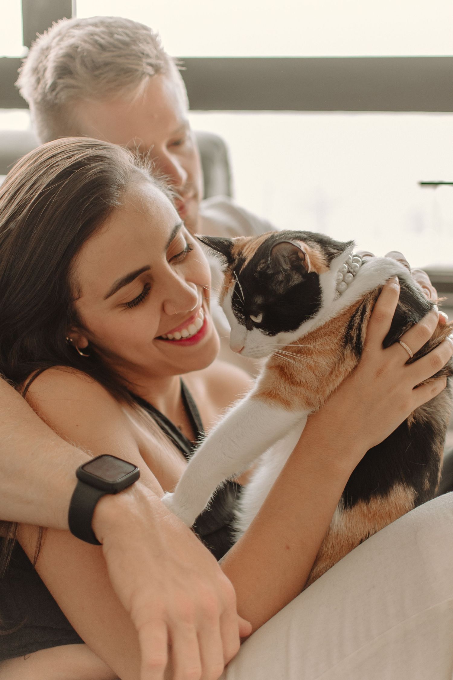 Couple relaxes on couch with their calico cat, sharing a tender moment in natural light.