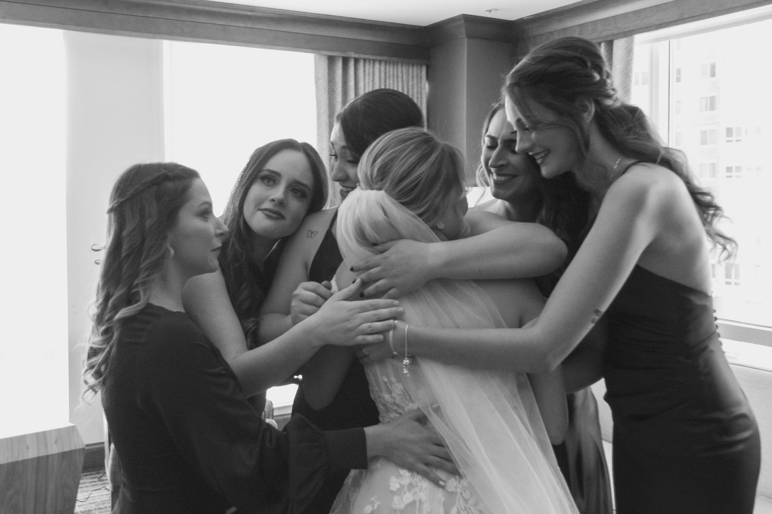Intimate black and white photo of bridesmaids in dark dresses sharing an emotional group hug with the bride.