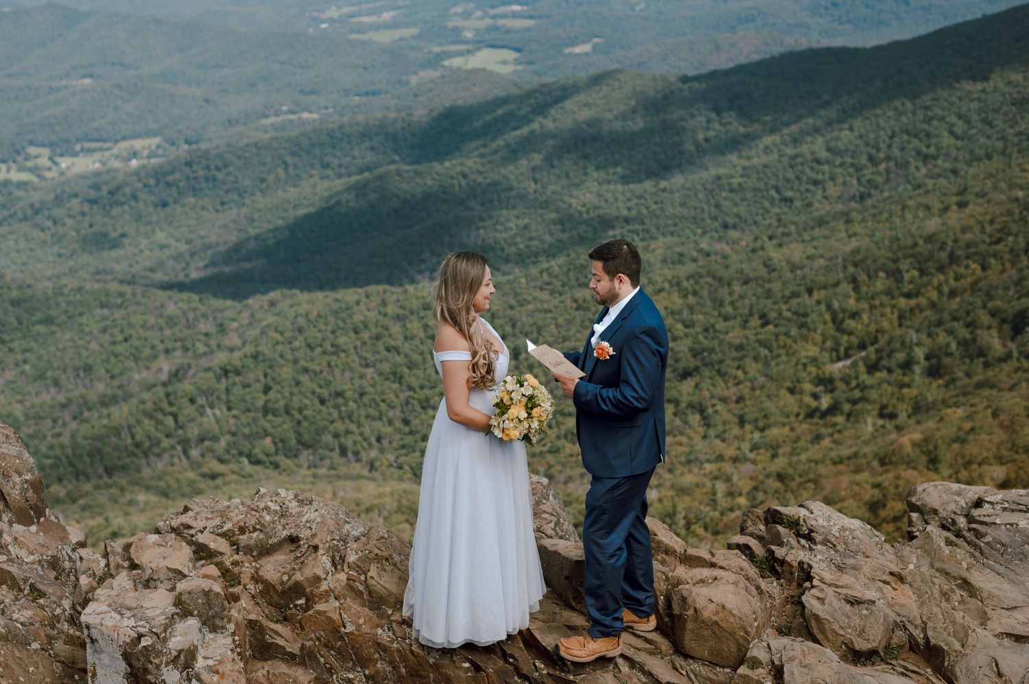 A couple in wedding attire stands on a mountain summit overlooking a scenic valley landscape on their special day.