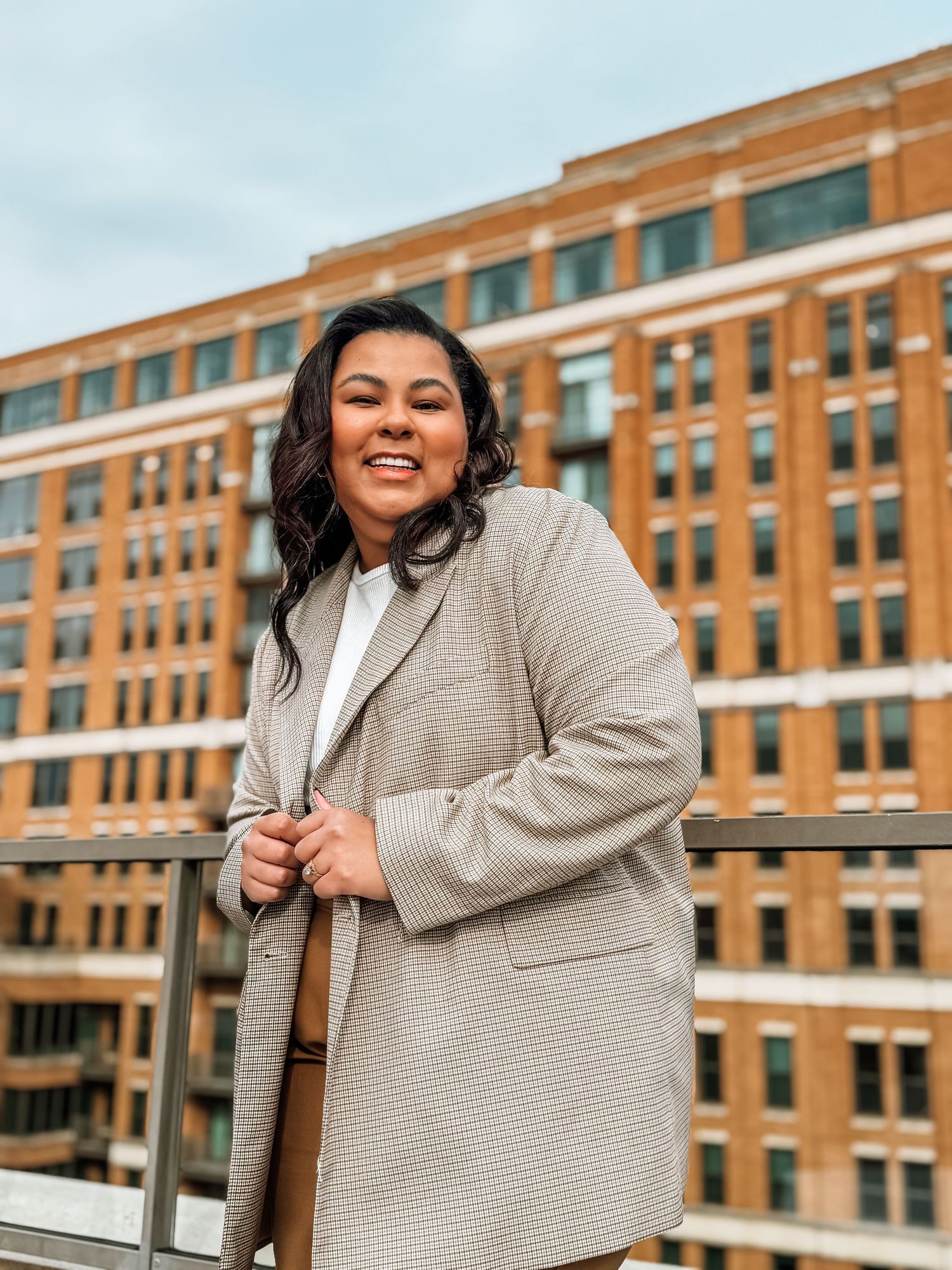 Someone wearing a cozy beige sweater coat poses outdoors against a brick apartment building on an overcast day.