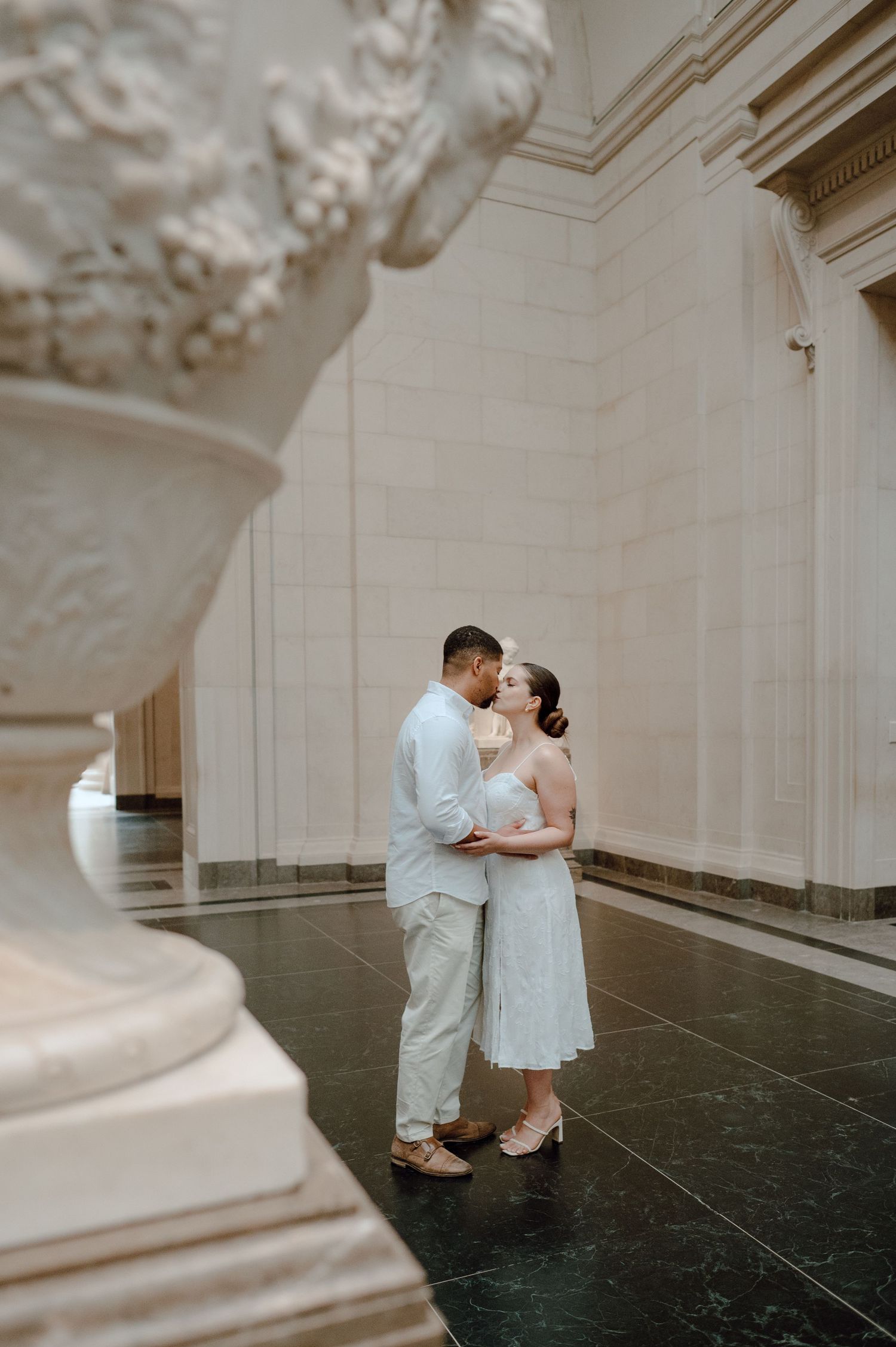 A romantic couple embraces in an elegant hallway with white marble columns and ornate architectural details.