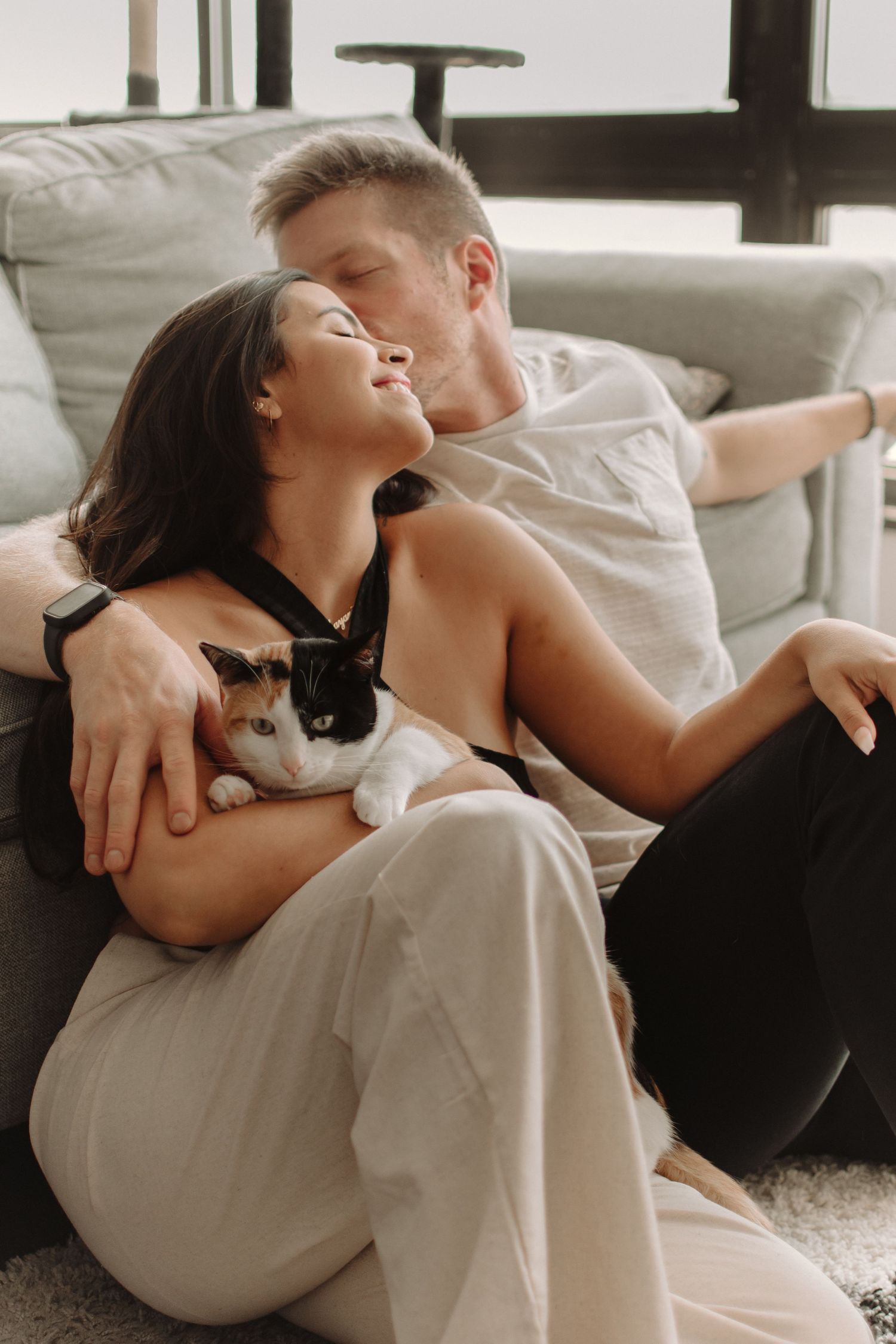 A couple relaxes on a couch with their small black and white cat in a warmly lit living room.