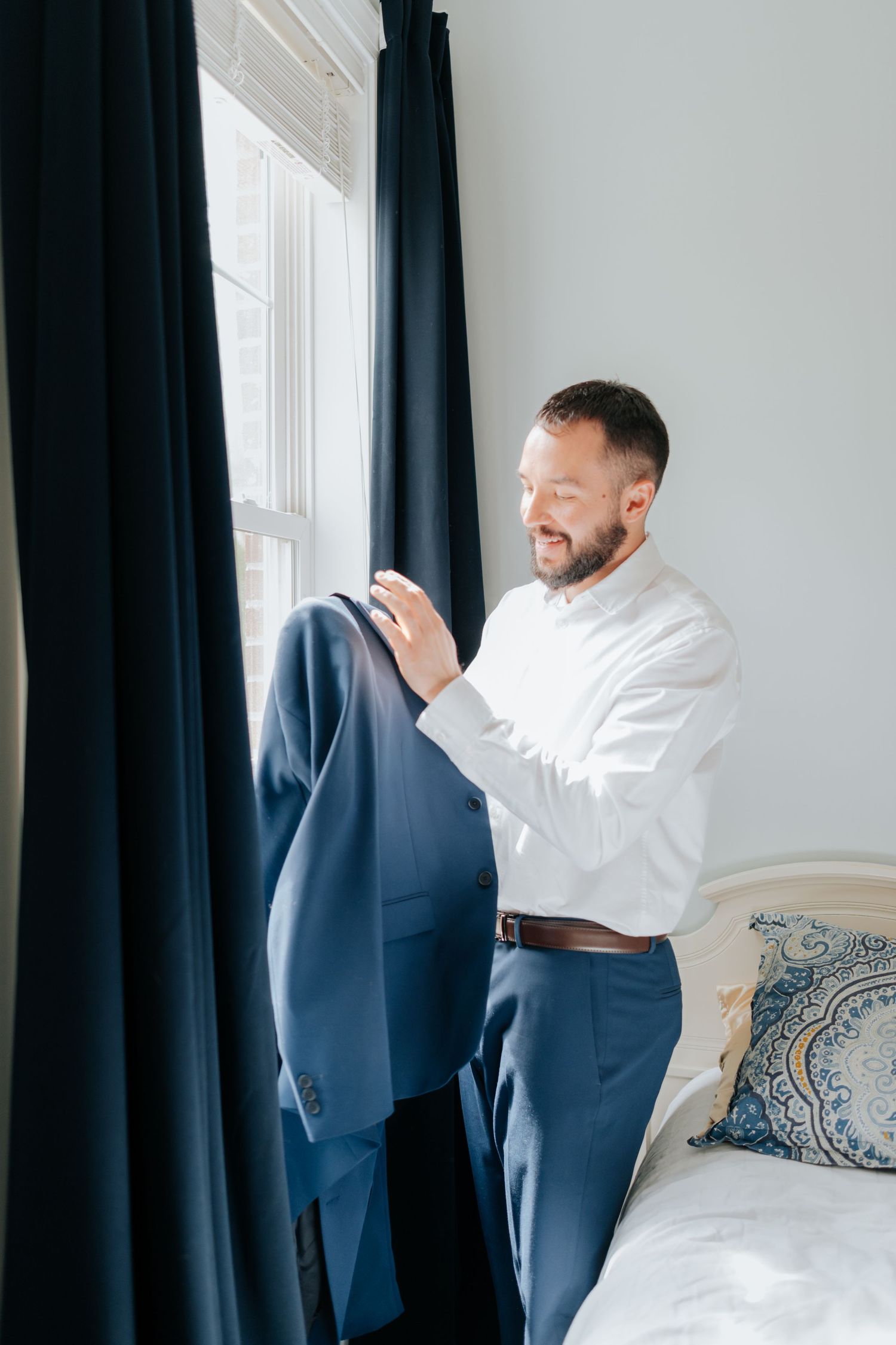 A person in formal attire carefully inspecting a navy blue suit jacket by a window with dark curtains.