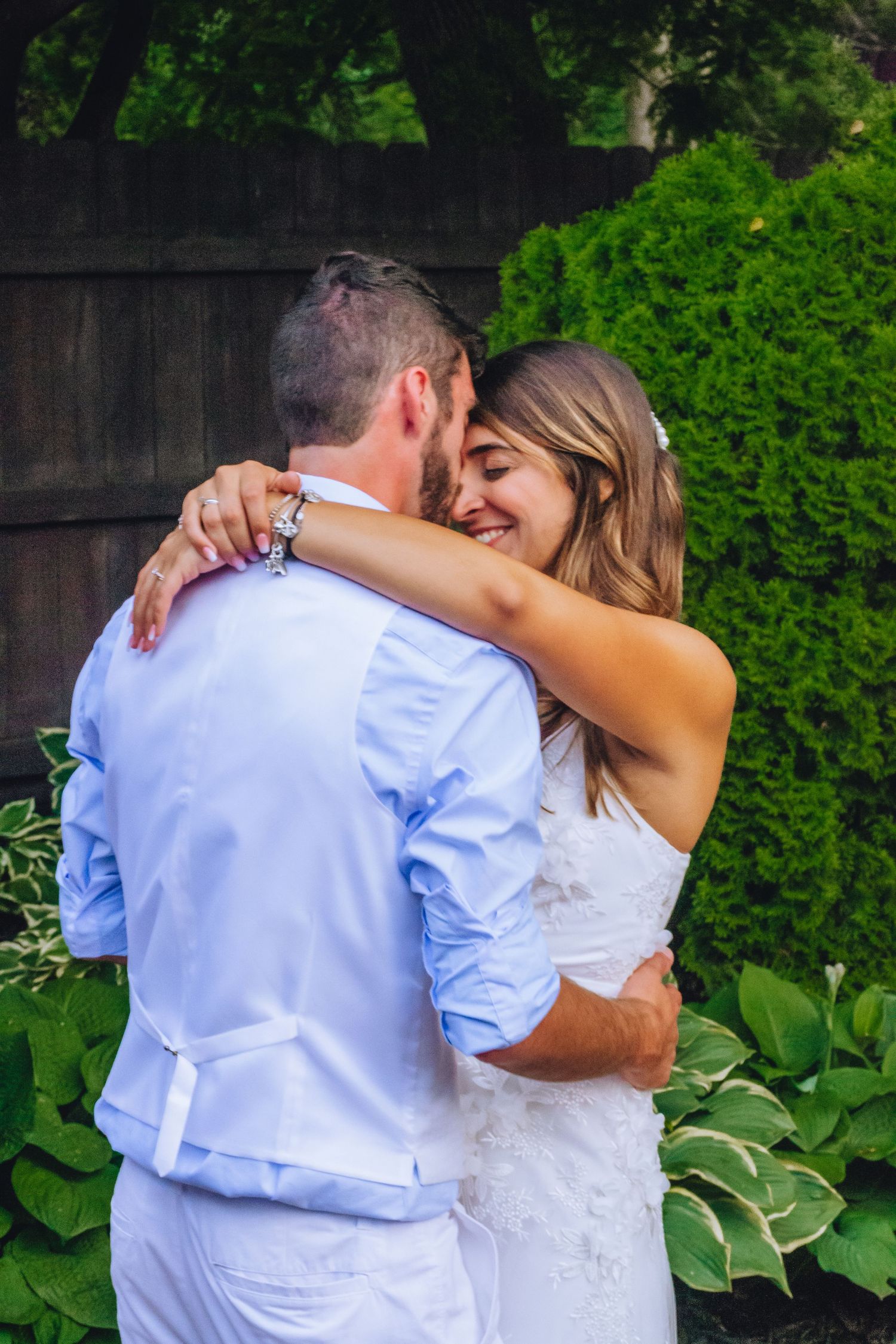 A couple embraces joyfully in their wedding attire against a lush green garden backdrop with blooming flowers.
