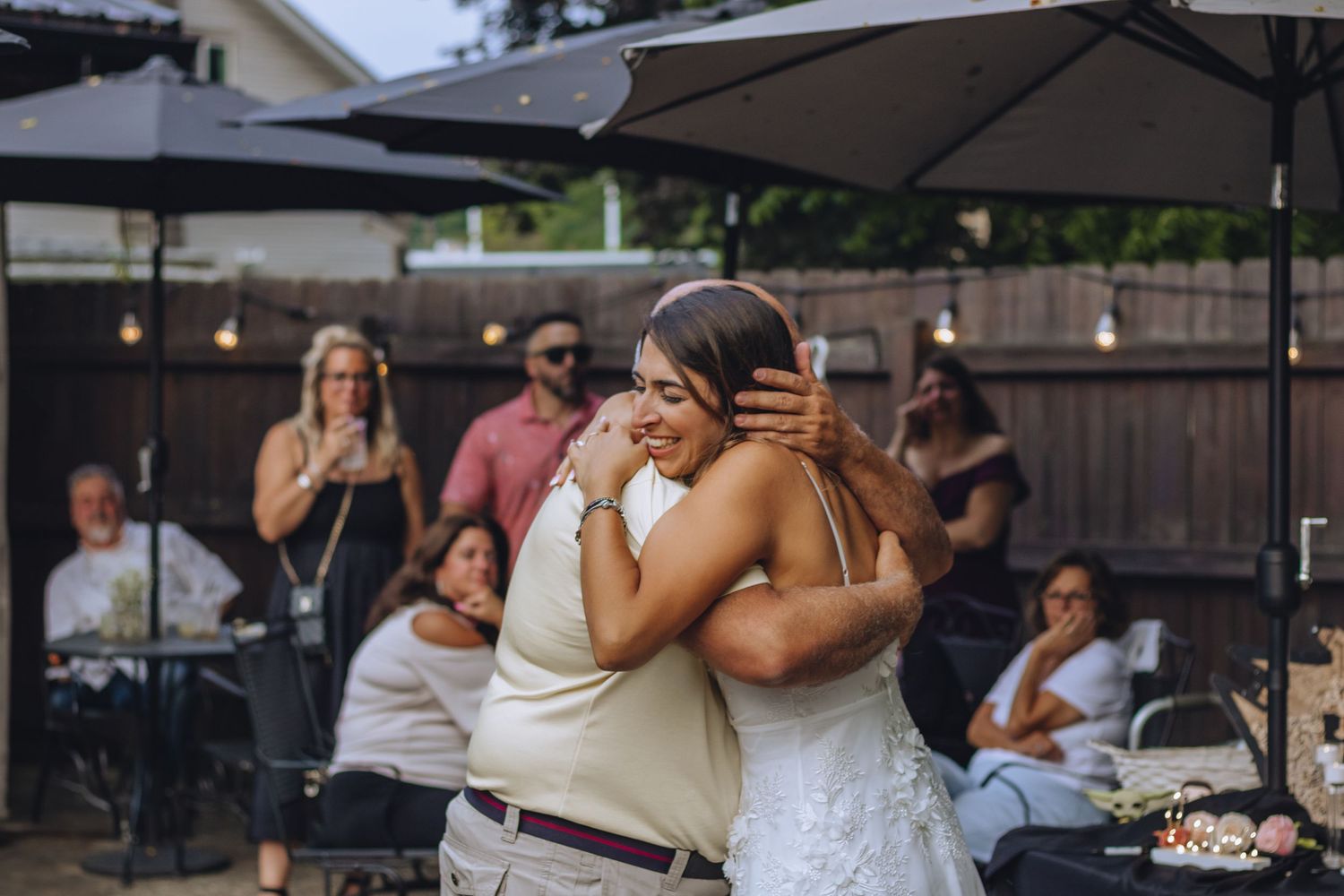 Two people share an emotional embrace on a patio while party guests sit at tables in the background.