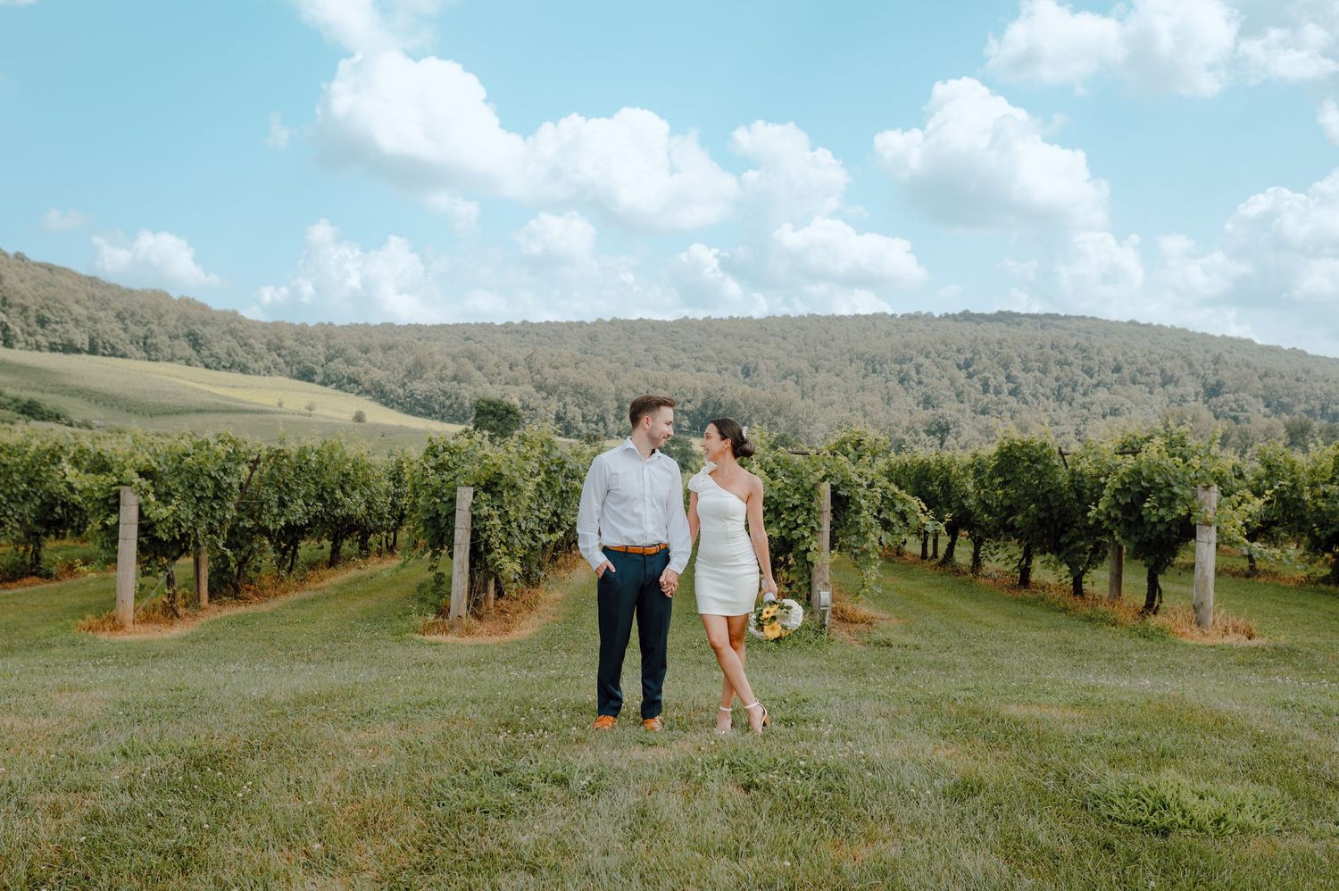 A couple walks hand in hand through a scenic vineyard with rolling hills and blue skies in the background.