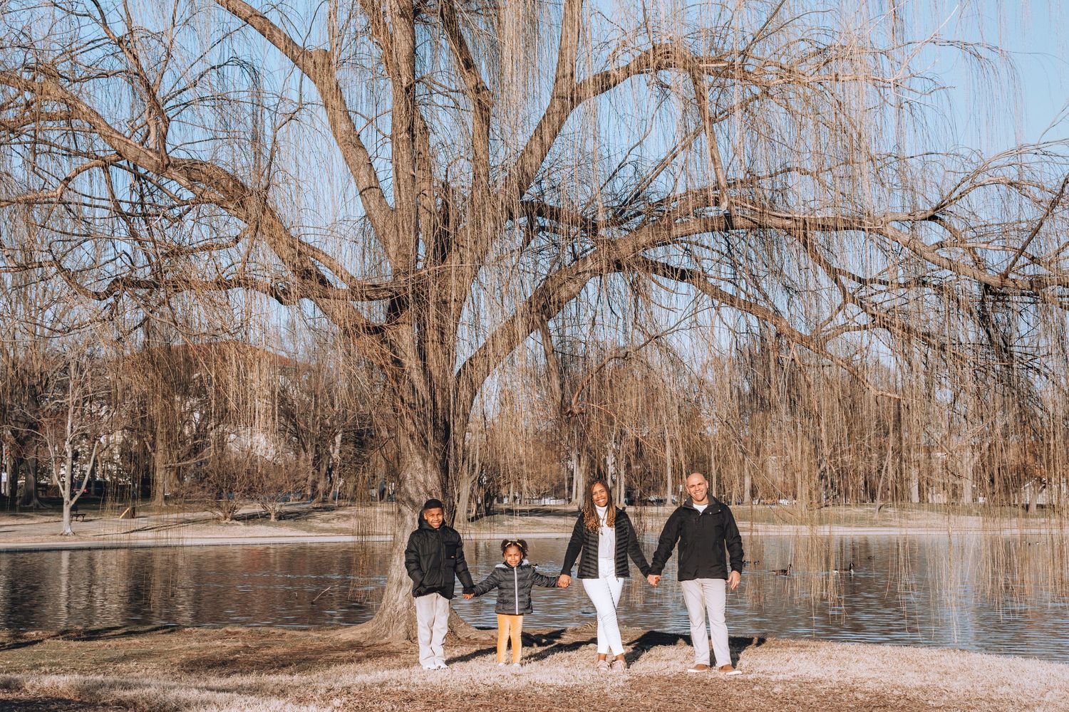 Family standing together by lake shore with bare winter trees in background.