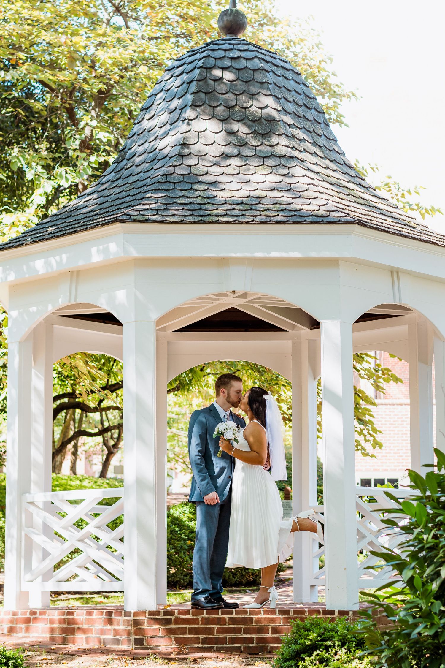 Newlyweds share a tender moment in a white wooden gazebo with ornate shingle roof and brick base in a garden setting.