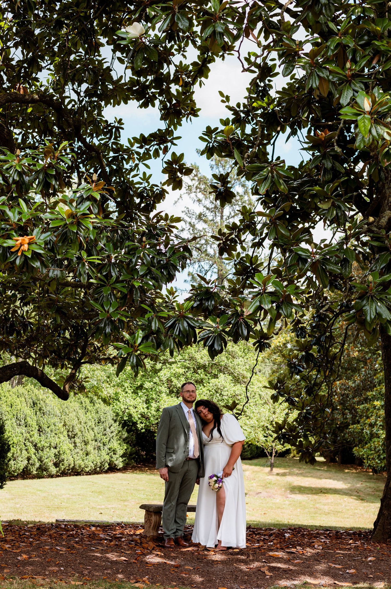 A couple stands together under a lush magnolia tree in an outdoor garden setting on a sunny day.