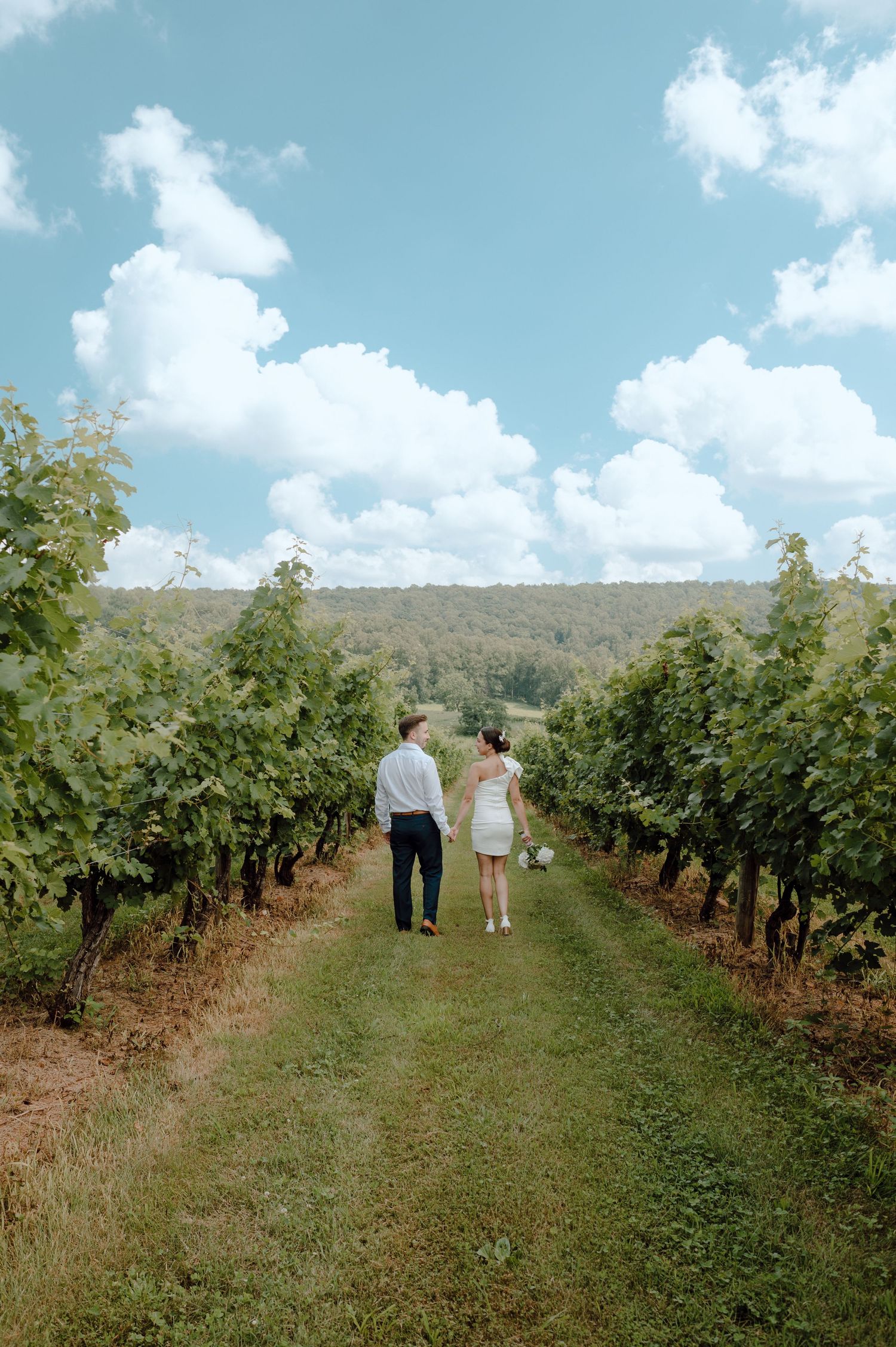 A couple strolls hand in hand through lush green vineyard rows on a beautiful summer day with blue skies overhead.