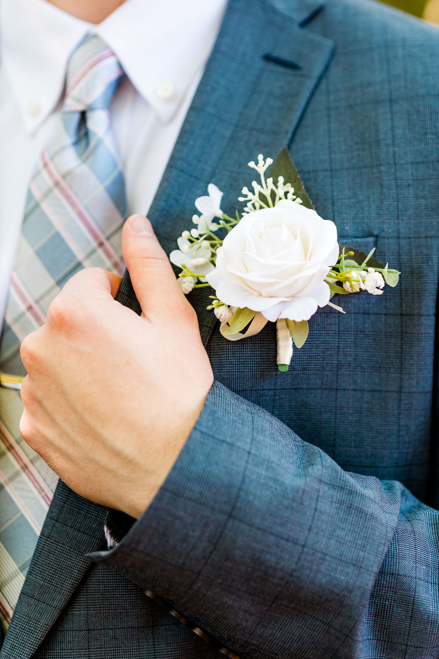 A white rose boutonniere with delicate greenery is pinned to a navy suit jacket with a plaid tie.