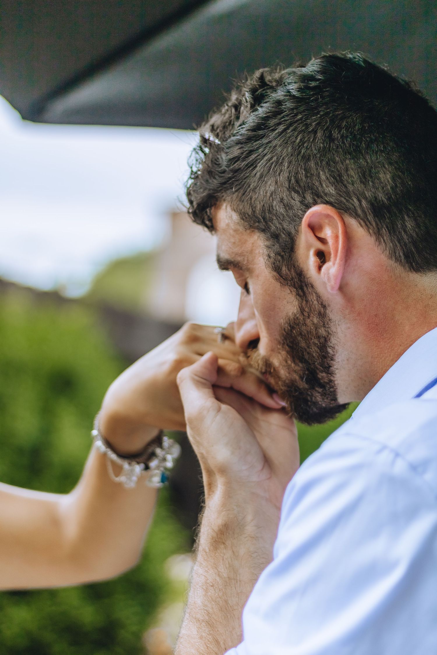 A groom in a white shirt kisses his bride's hand during a wedding ceremony outdoors.