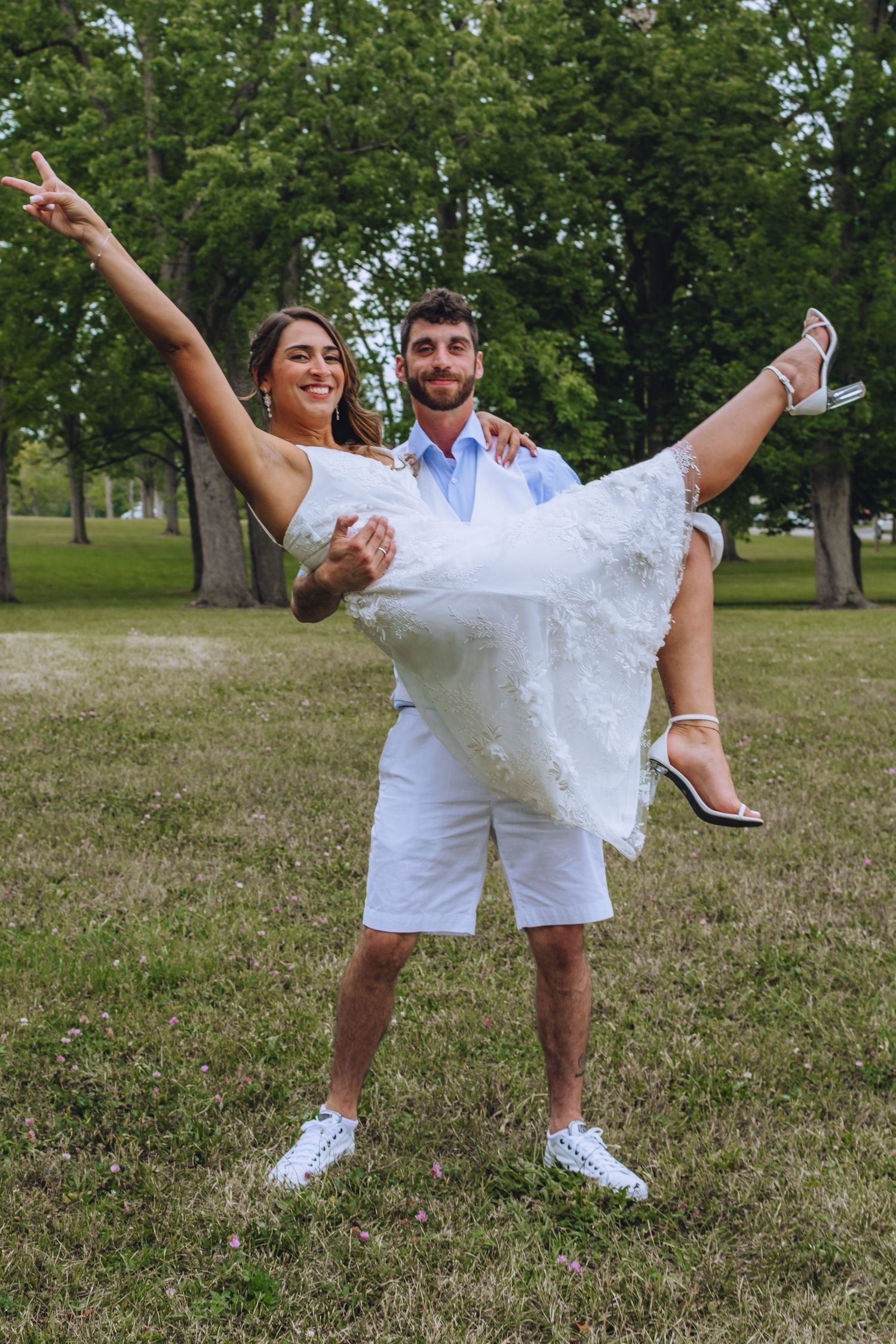 A joyful couple poses in a park, with the bride wearing a white dress being carried by her partner in white shorts.