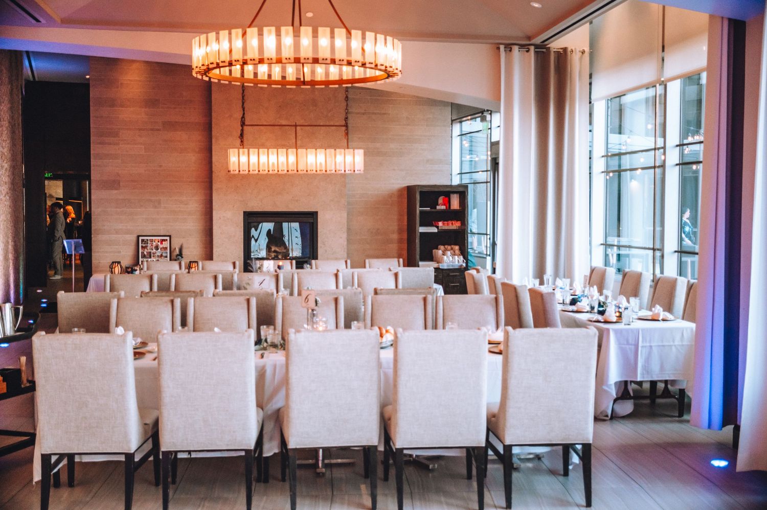 Modern dining room with white chairs, wooden walls, and an elegant circular chandelier in an upscale restaurant setting.