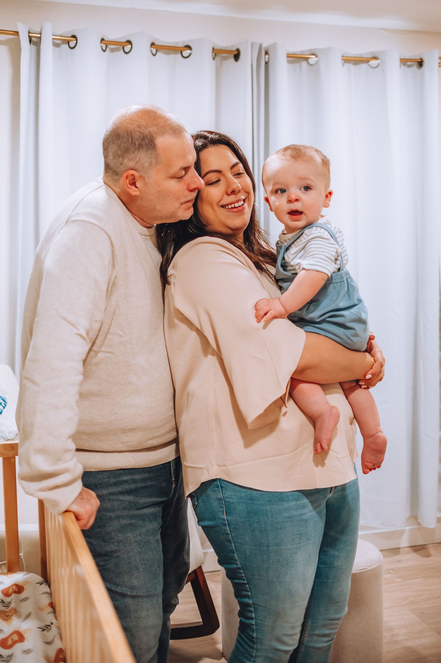 Intimate family portrait in a nursery with white curtains and wooden furniture.