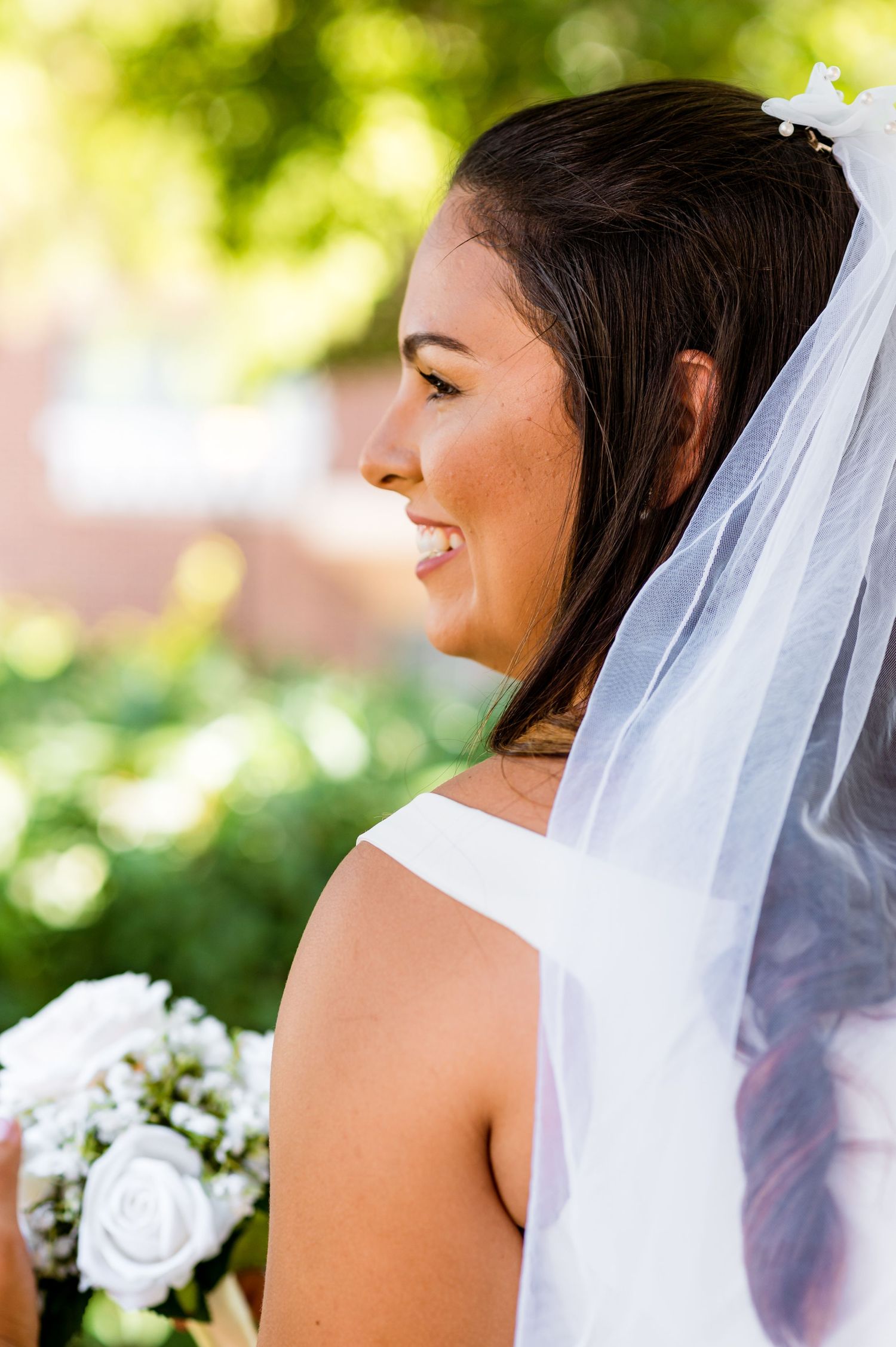 Side profile of bride wearing veil and holding white bouquet with natural bokeh background.