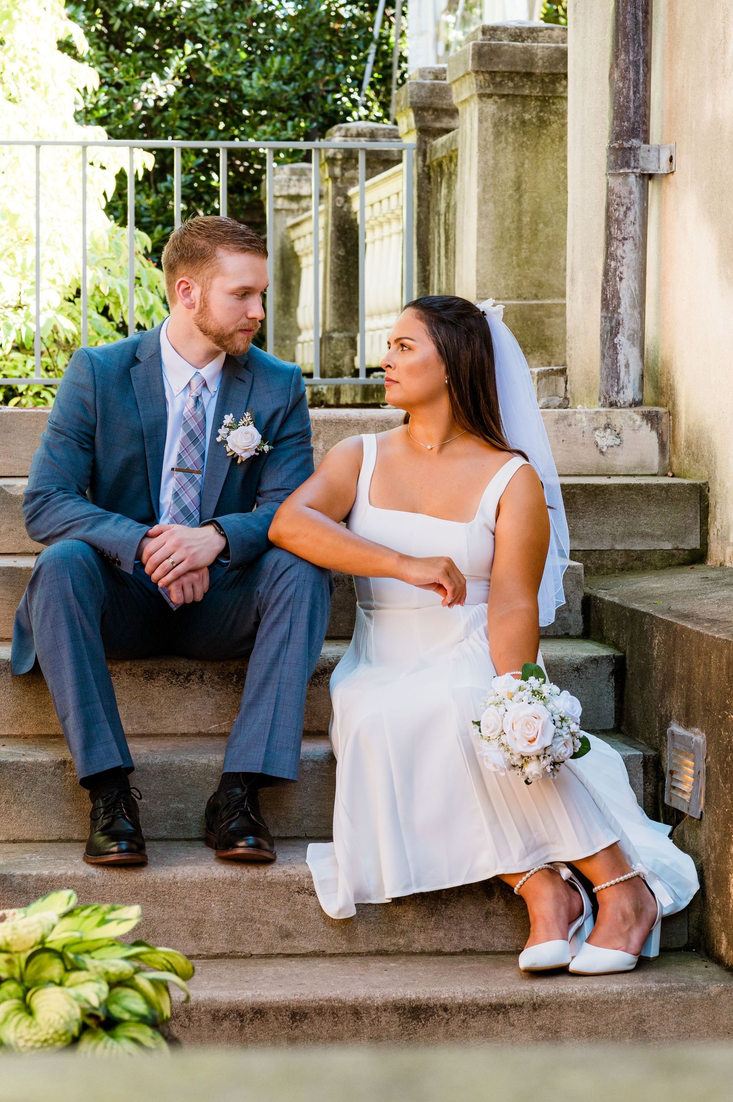 Newlywed couple sitting on stone steps in elegant wedding attire with bouquet.
