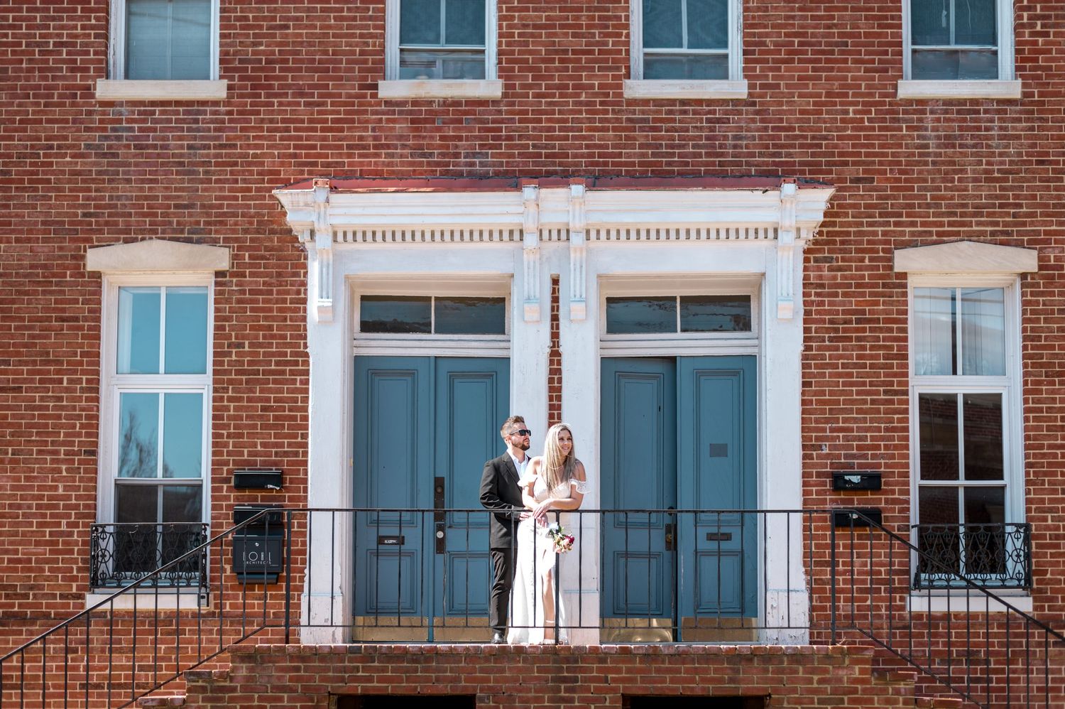 Wedding couple stands in front of historic brick building with symmetrical blue doors.