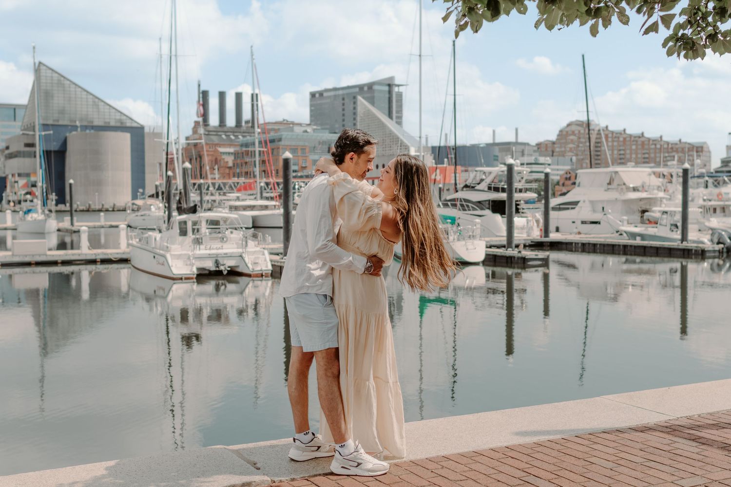 Couple embraces on a wooden dock with boats and industrial buildings in the background at a marina.