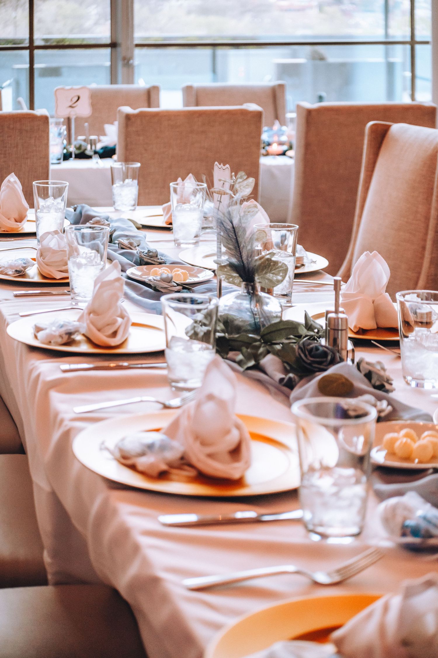 Elegant table settings at a banquet with peach tablecloths, white napkins and greenery decorations in a bright space.