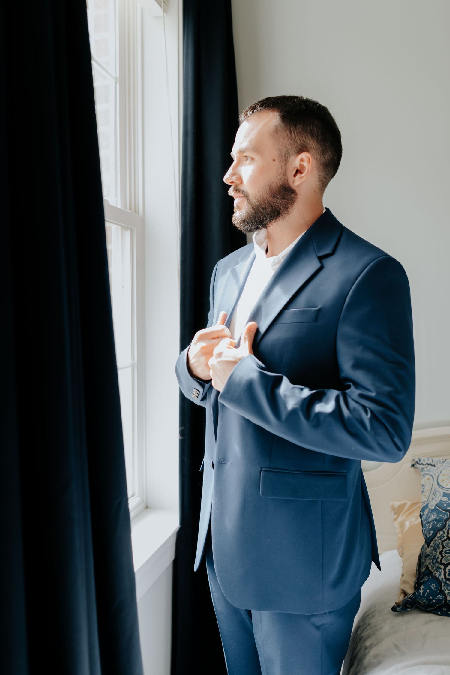 Person in a navy blue suit adjusts their tie while looking thoughtfully out a window with dark curtains.