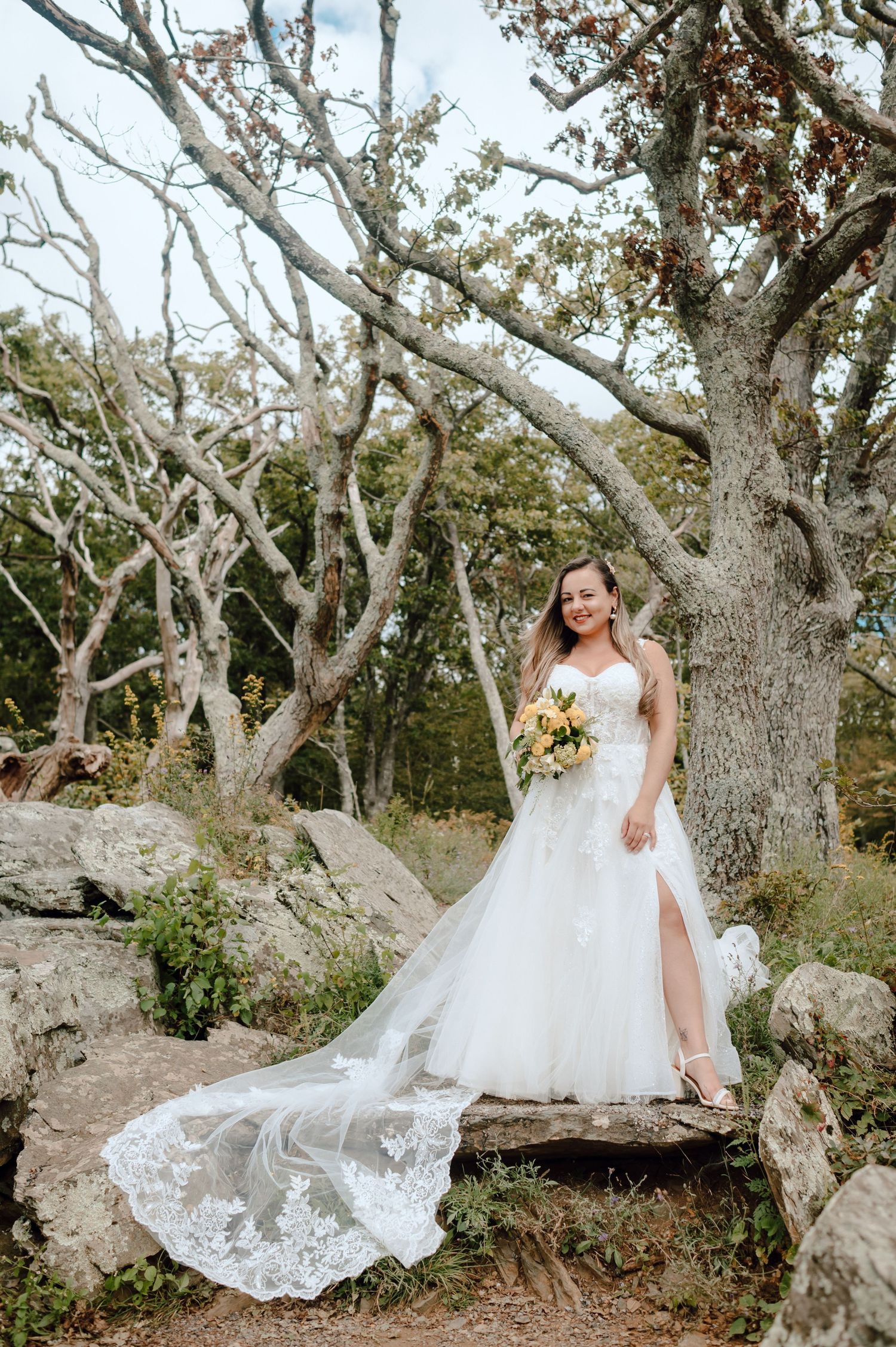 Bride in flowing white lace gown poses gracefully among twisted oak trees in a rustic outdoor setting.