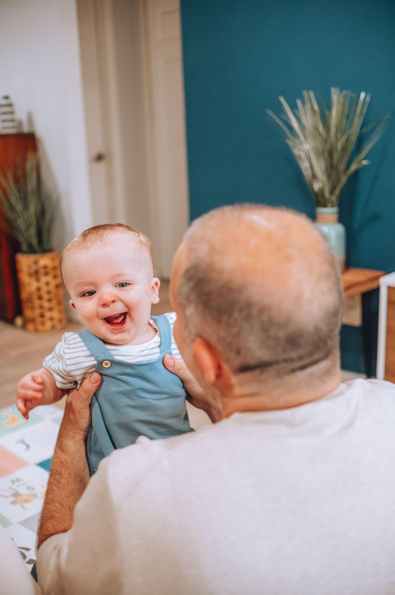 Baby in blue overalls laughs joyfully during playtime on floor.