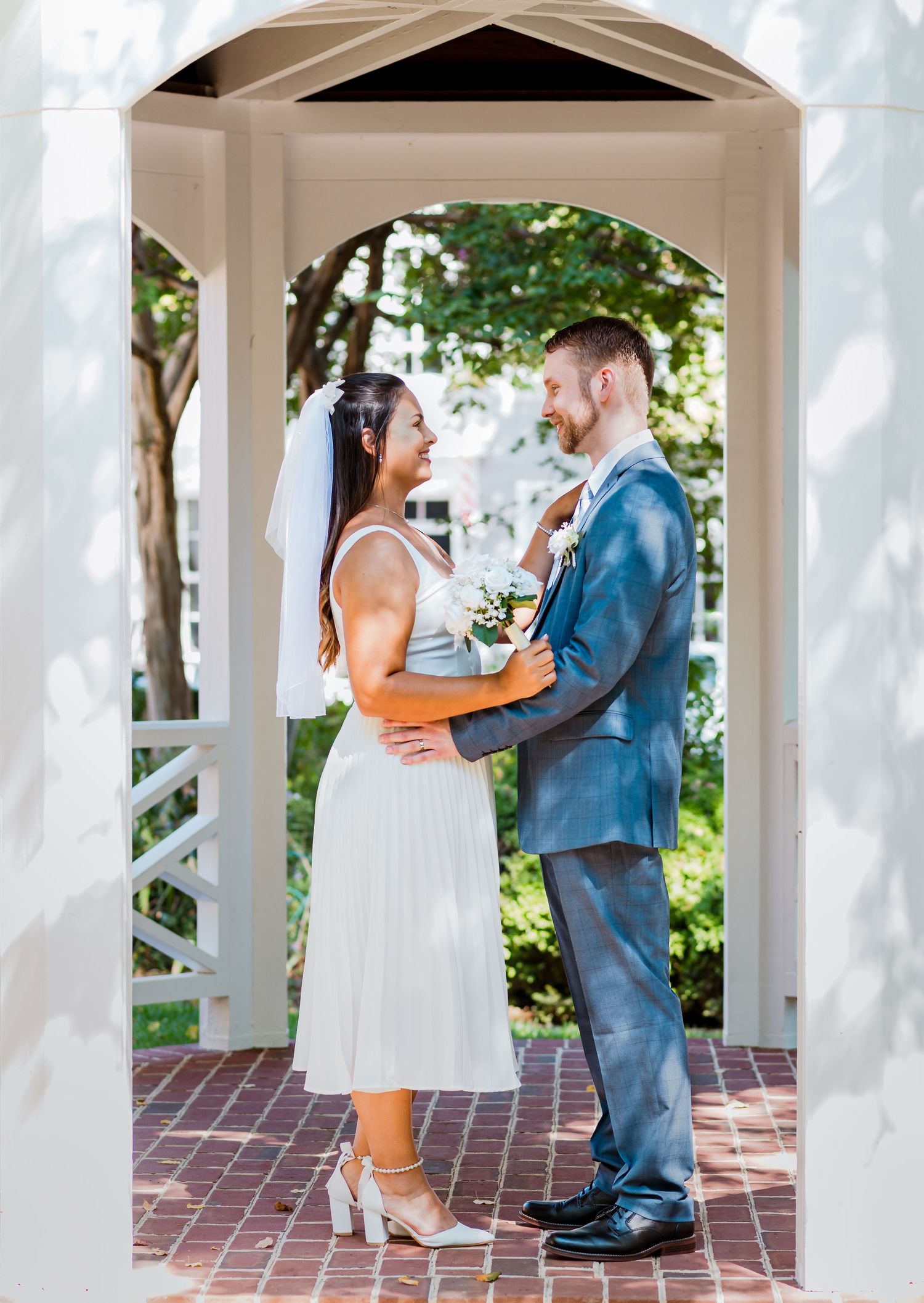 Wedding couple sharing intimate moment under white garden gazebo on brick path.