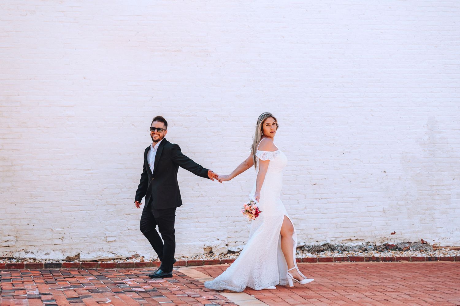 A couple holds hands while walking along a brick wall in wedding attire.