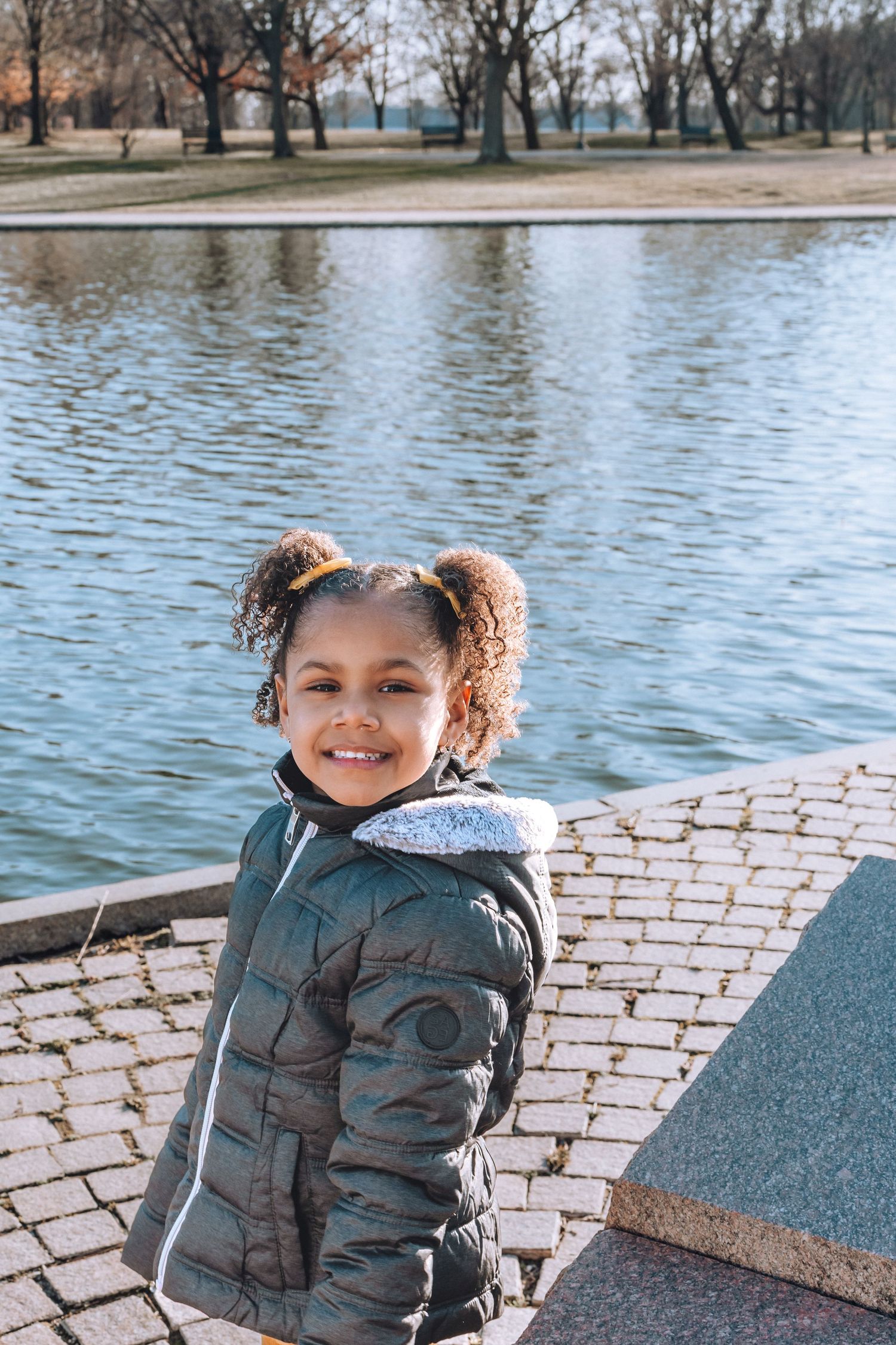 Child in winter coat smiling by water on brick pathway.