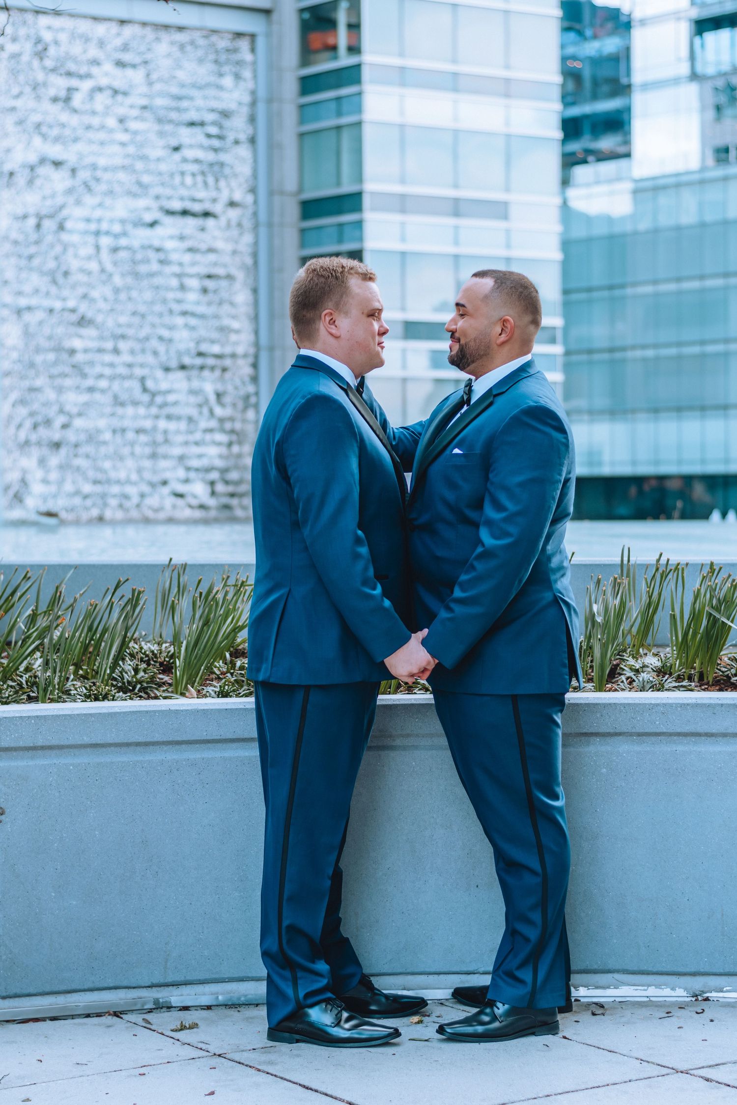 Two people in navy blue suits share an intimate moment in front of a modern building with concrete planters.