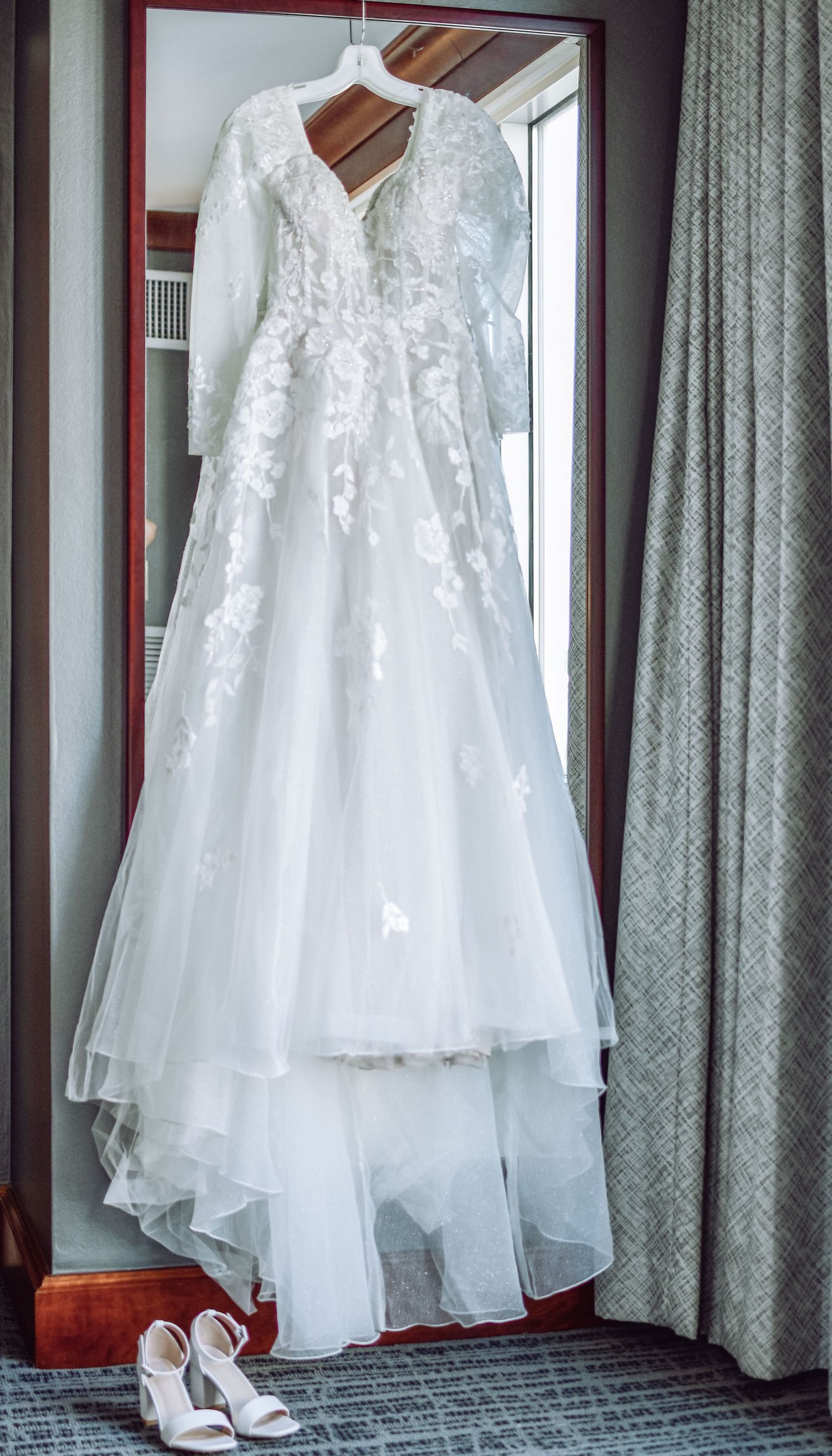 An elegant white lace wedding dress with long sleeves and flowing tulle skirt hangs by a window, with white heels below.