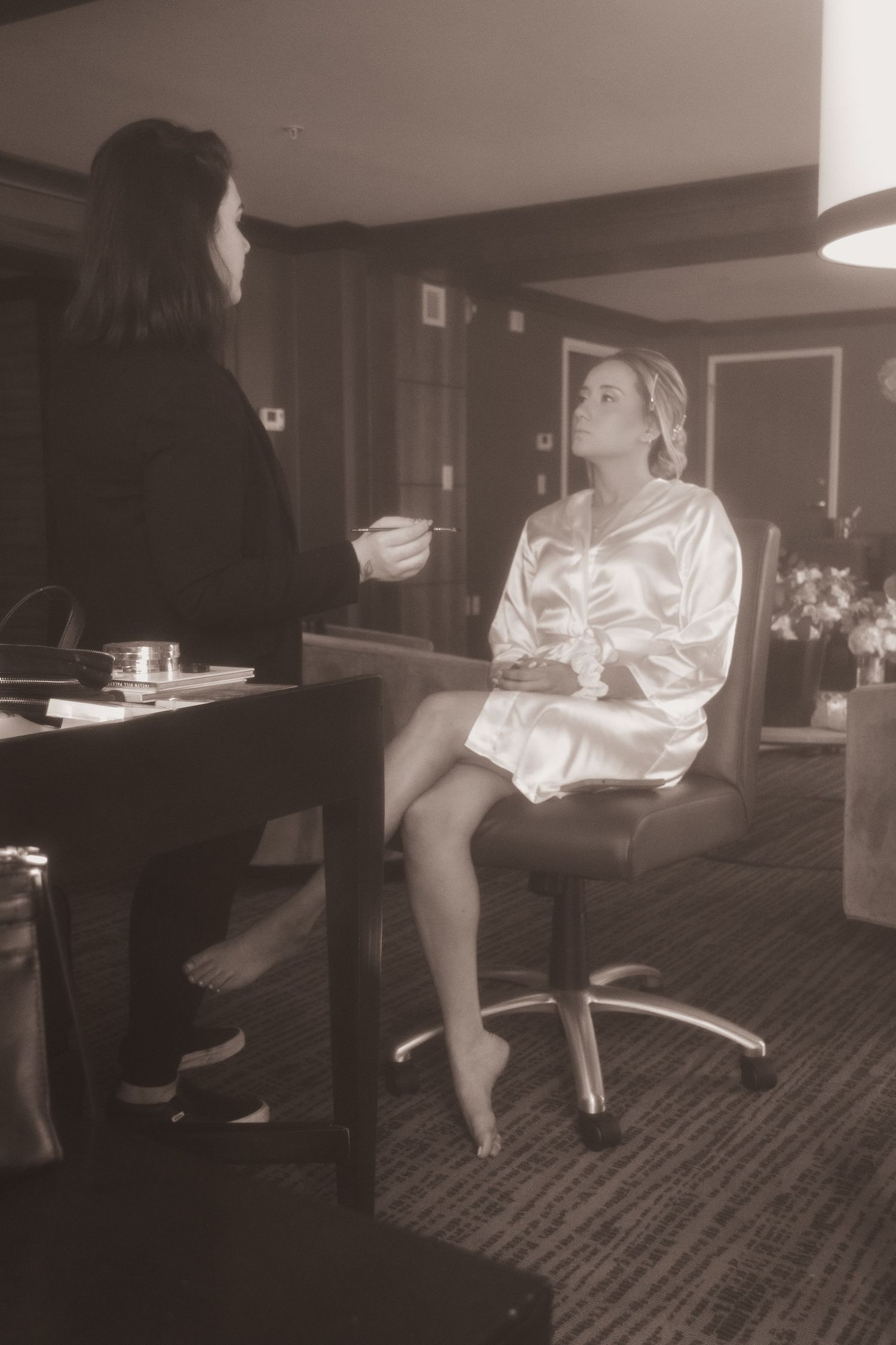 Behind-the-scenes black and white photo of makeup application in a hotel room showing someone in a silk robe getting ready.