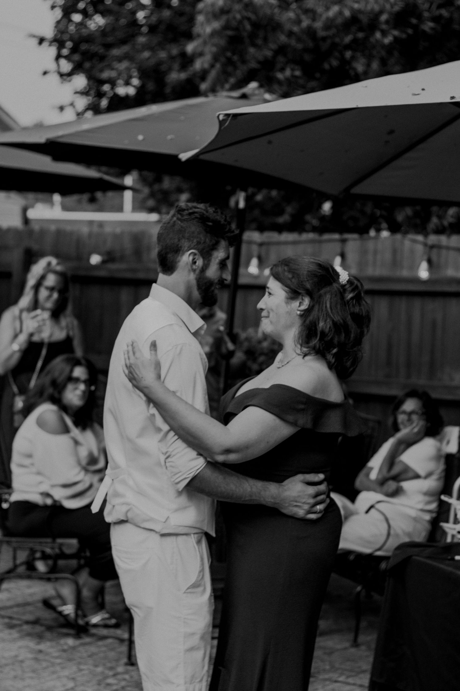 A couple dances together at an outdoor party under black umbrellas while guests look on in the background.