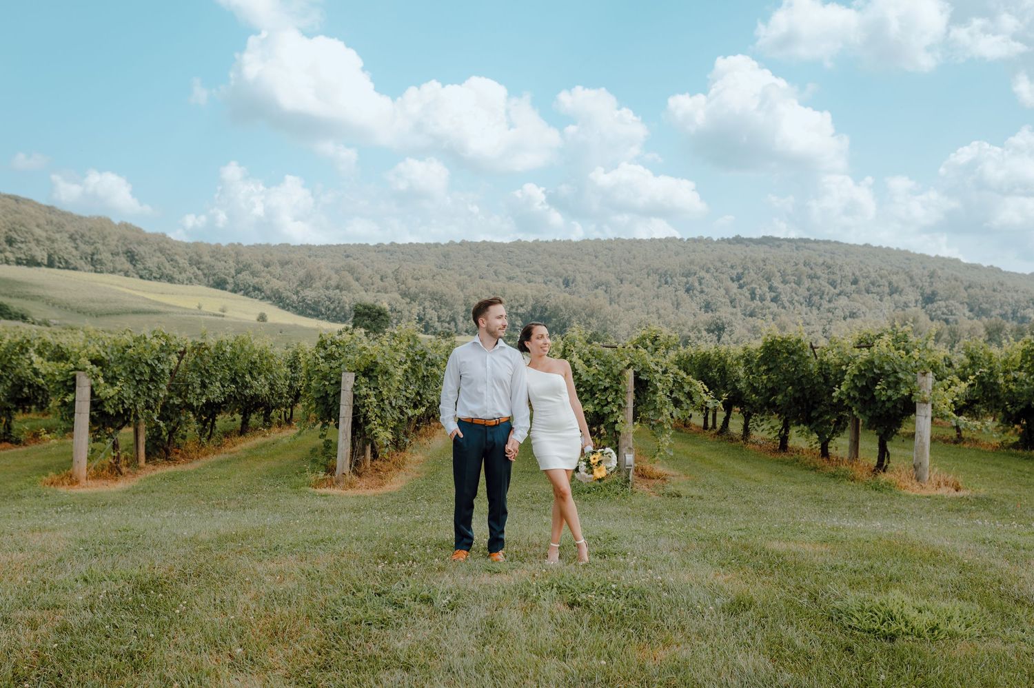 A couple stands together in a picturesque vineyard with rolling hills and blue skies in the background.