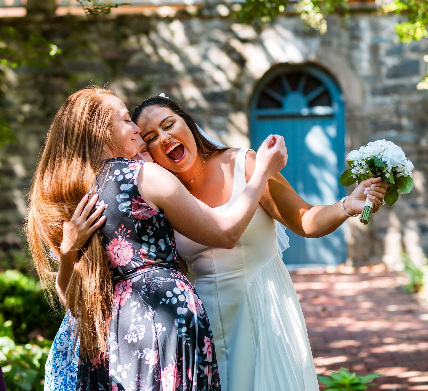 Joyful embrace between bride and guest in floral dress against stone building backdrop.