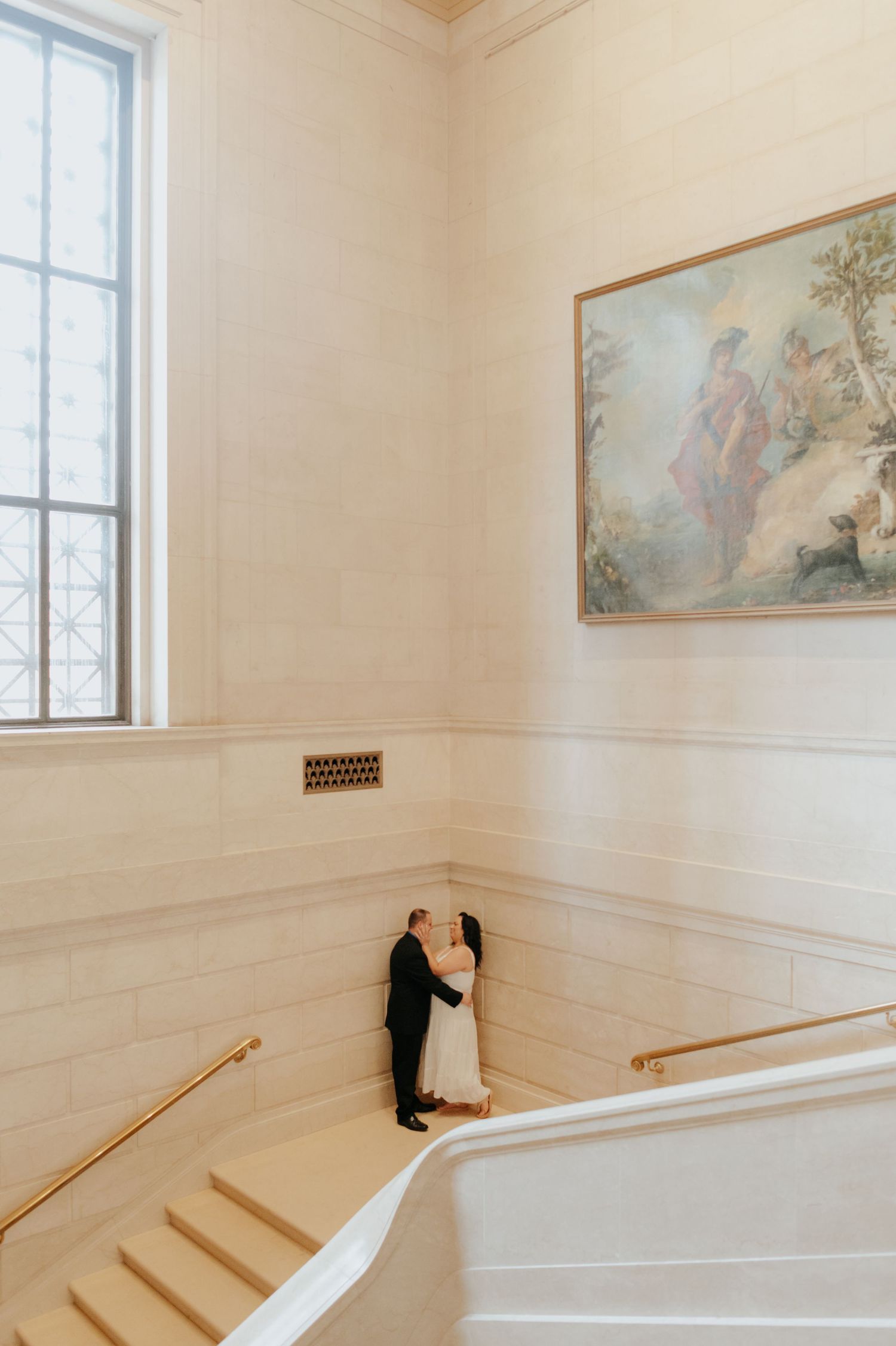Couple shares an intimate moment on an elegant white staircase landing with a large window and classical artwork.