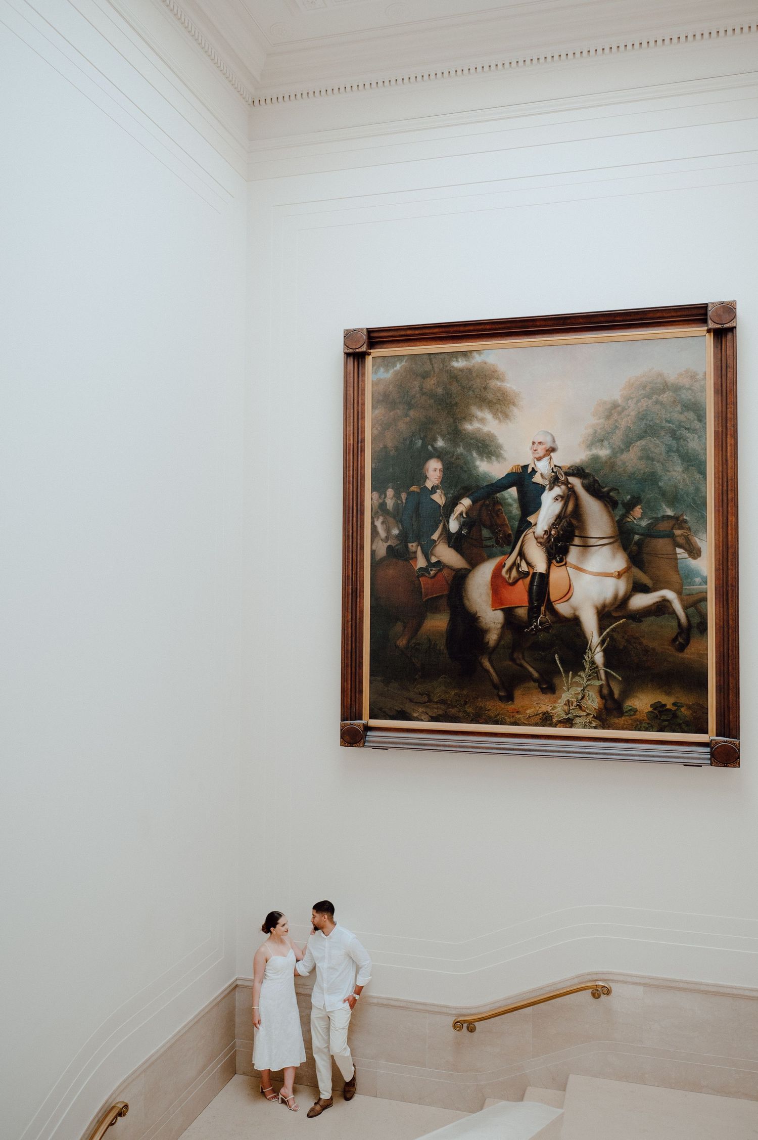 Two people stand on marble stairs beneath a grand classical equestrian painting in an elegant gallery setting.