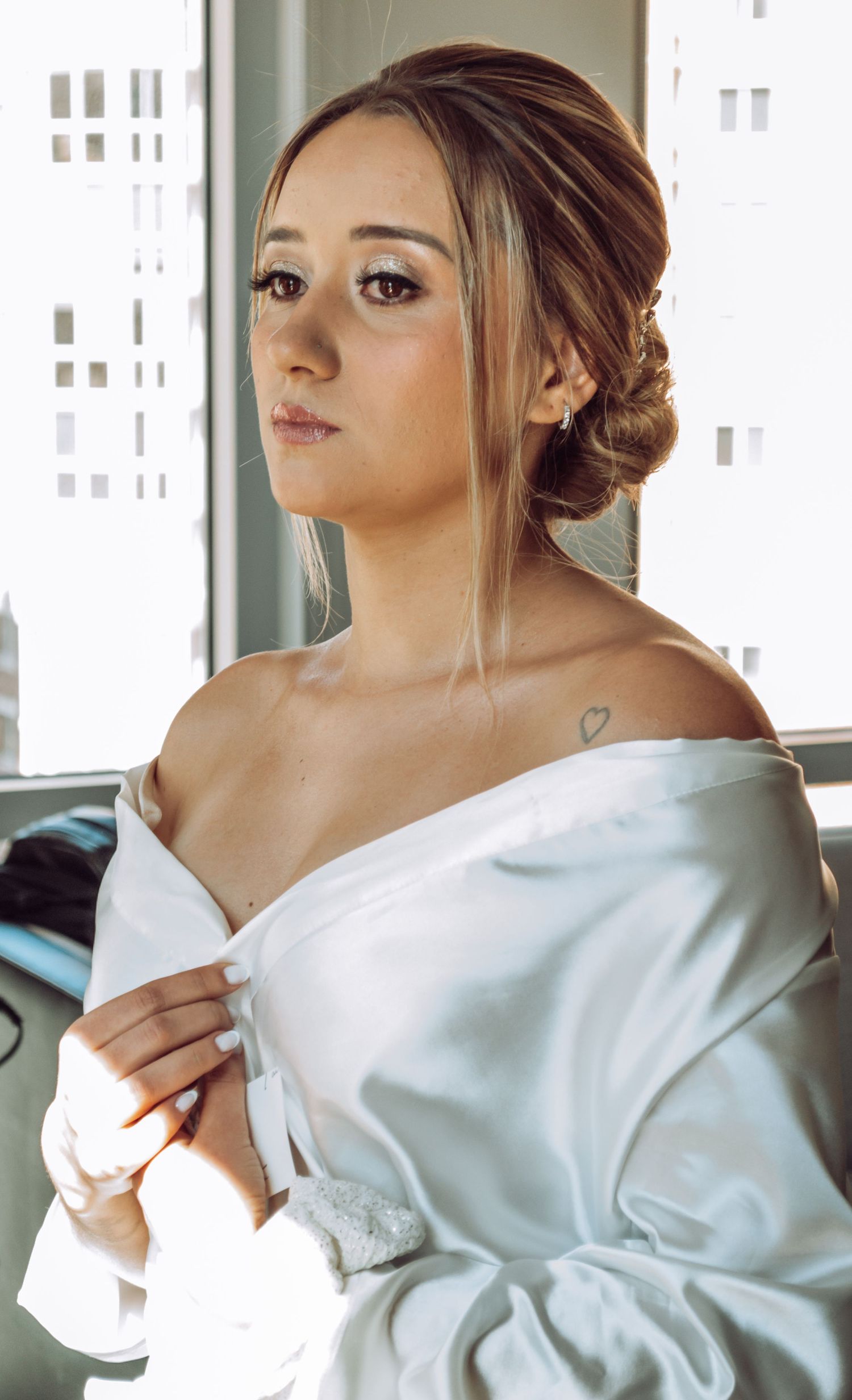 A bride in a white robe poses near a window with an elegant updo hairstyle and natural makeup.