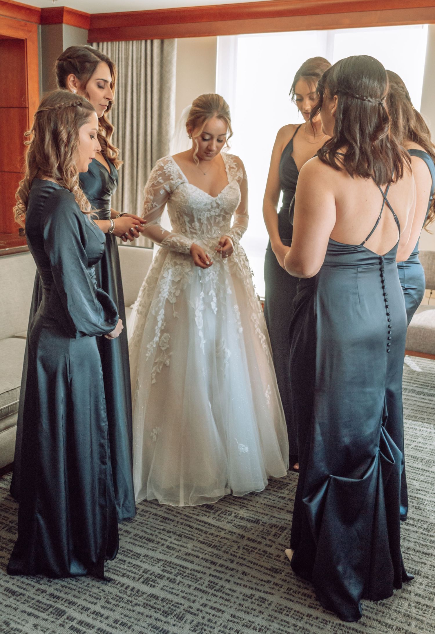 Bridesmaids in teal dresses help with wedding preparations in a hotel room while a white wedding gown hangs nearby.