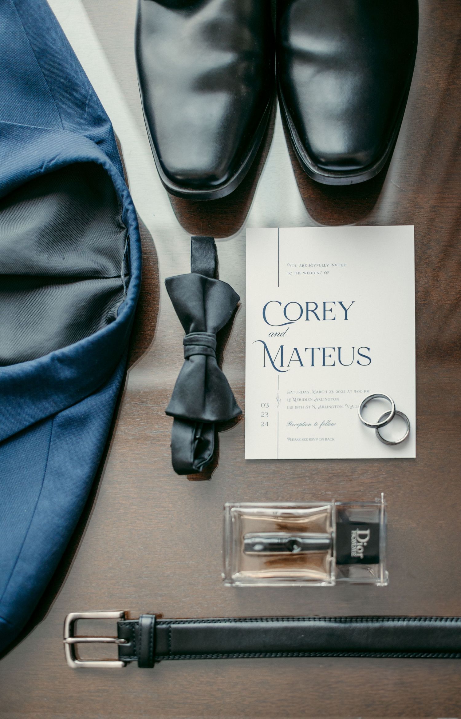 Wedding details featuring polished black dress shoes, blue suit jackets, leather belt and name cards on wooden surface.