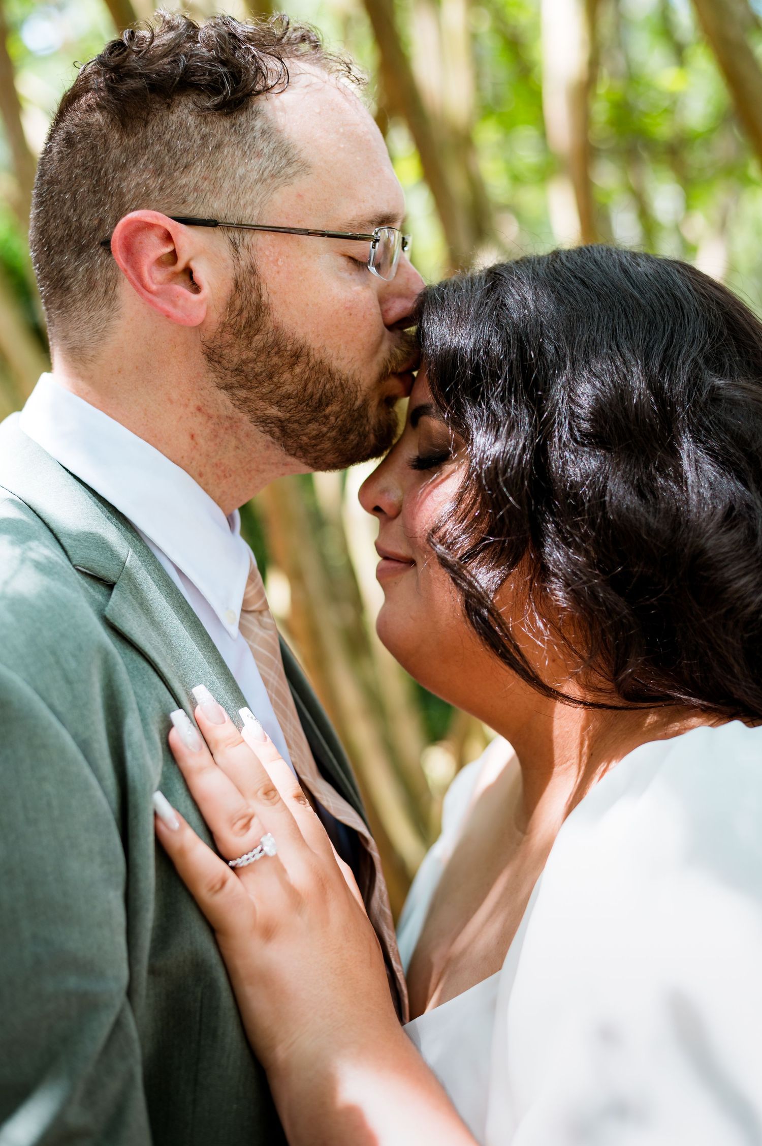 A romantic wedding couple shares an intimate kiss in a wooded outdoor setting.