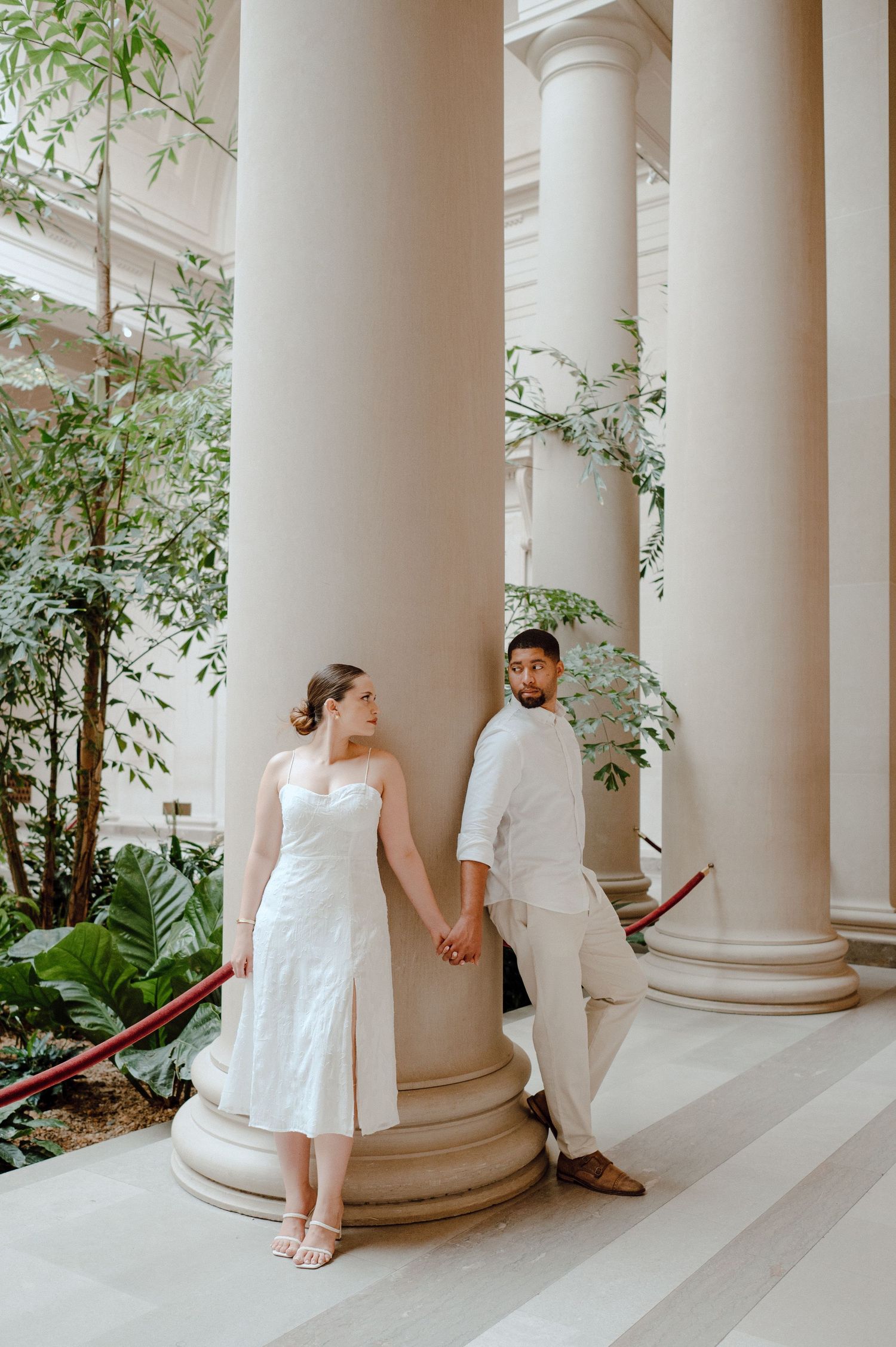 A couple poses romantically between classic white columns surrounded by indoor plants.