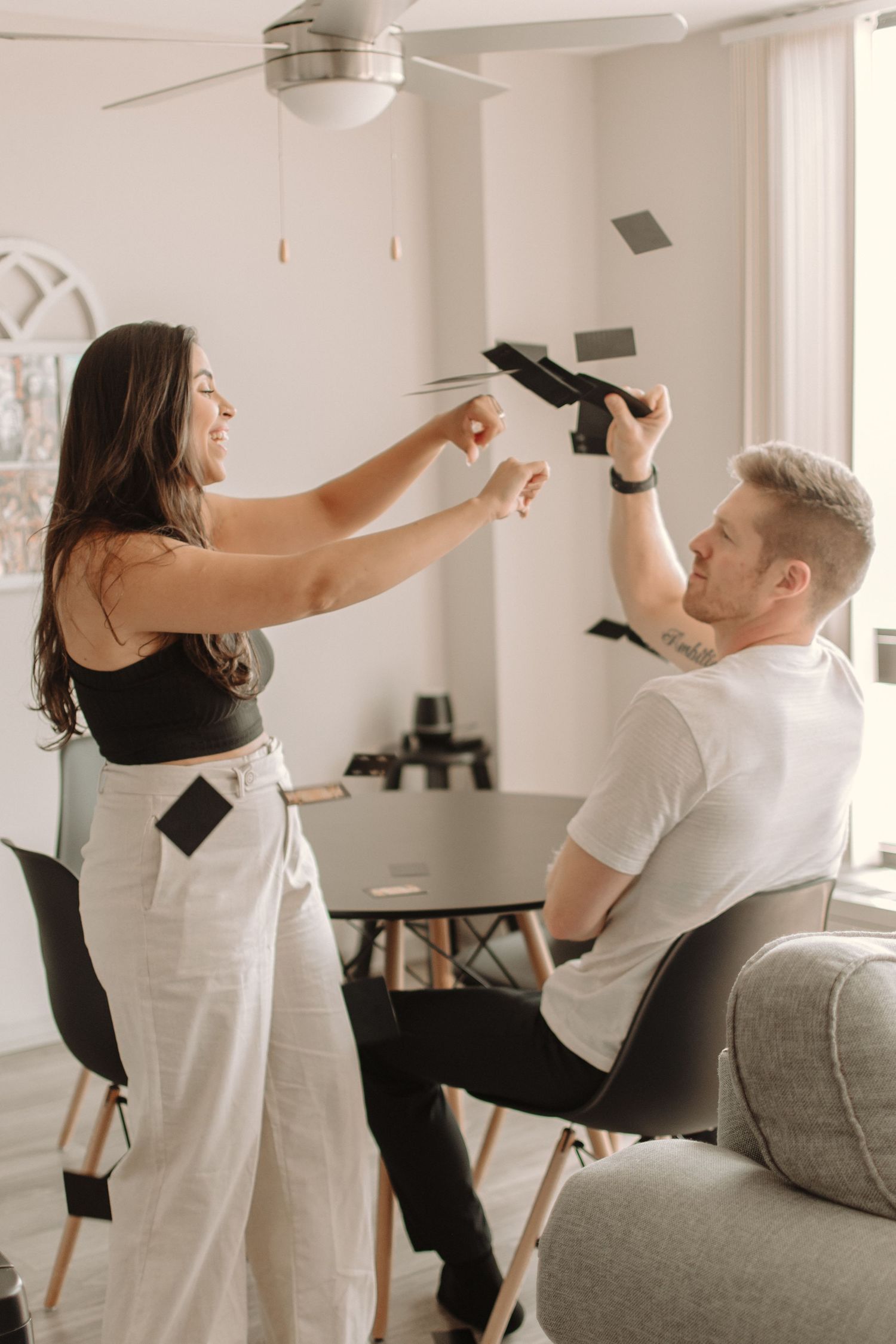 Playful couple having fun throwing paper cards at each other in a bright living room.