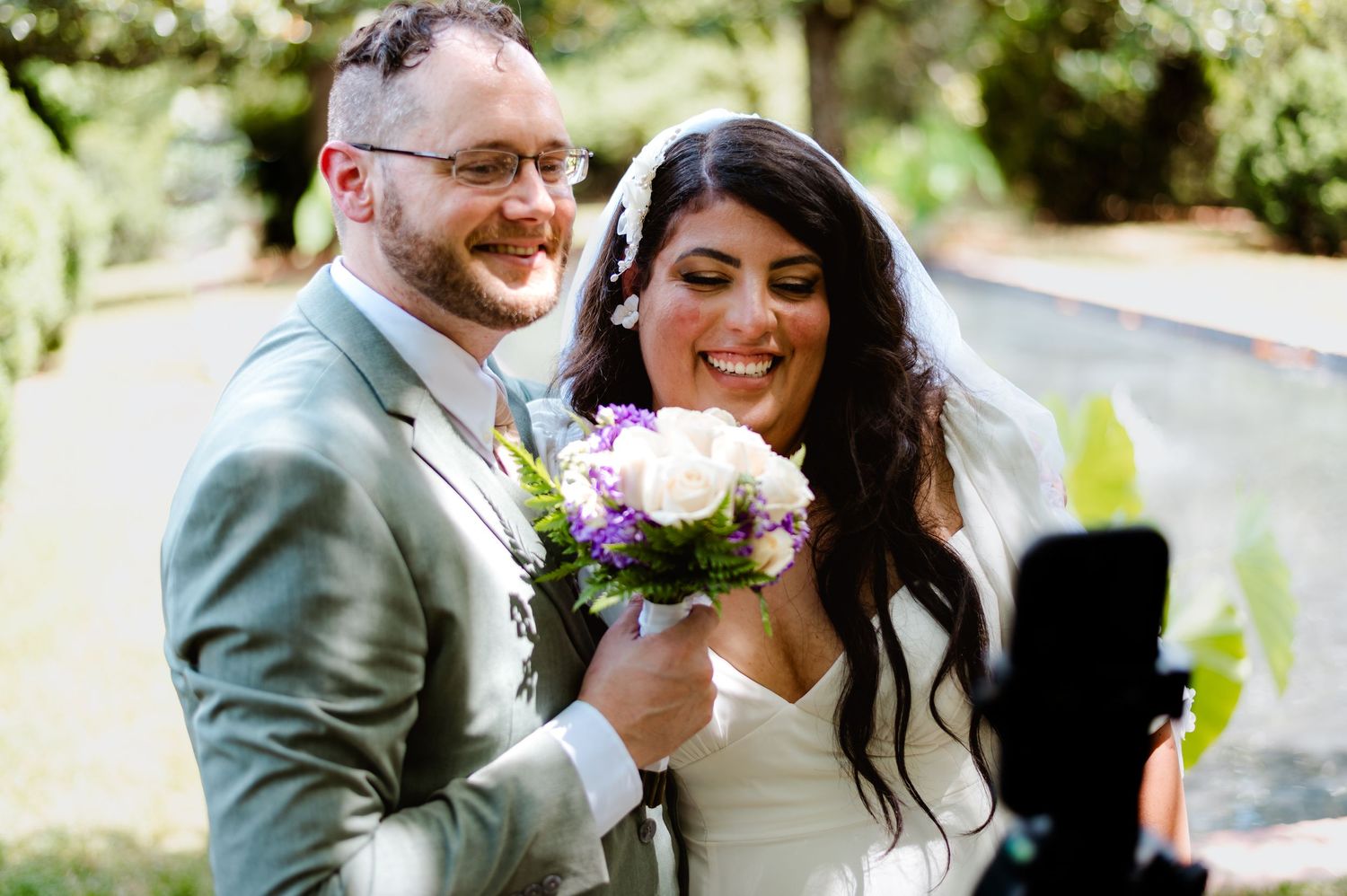 A joyful newlywed couple sharing a candid moment outdoors while holding a pink and white bouquet.