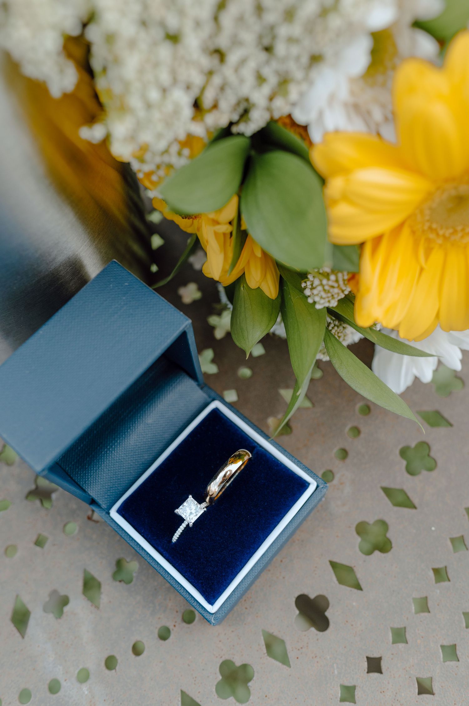 Diamond engagement ring in blue box with yellow sunflowers and white floral decorations on pale background.